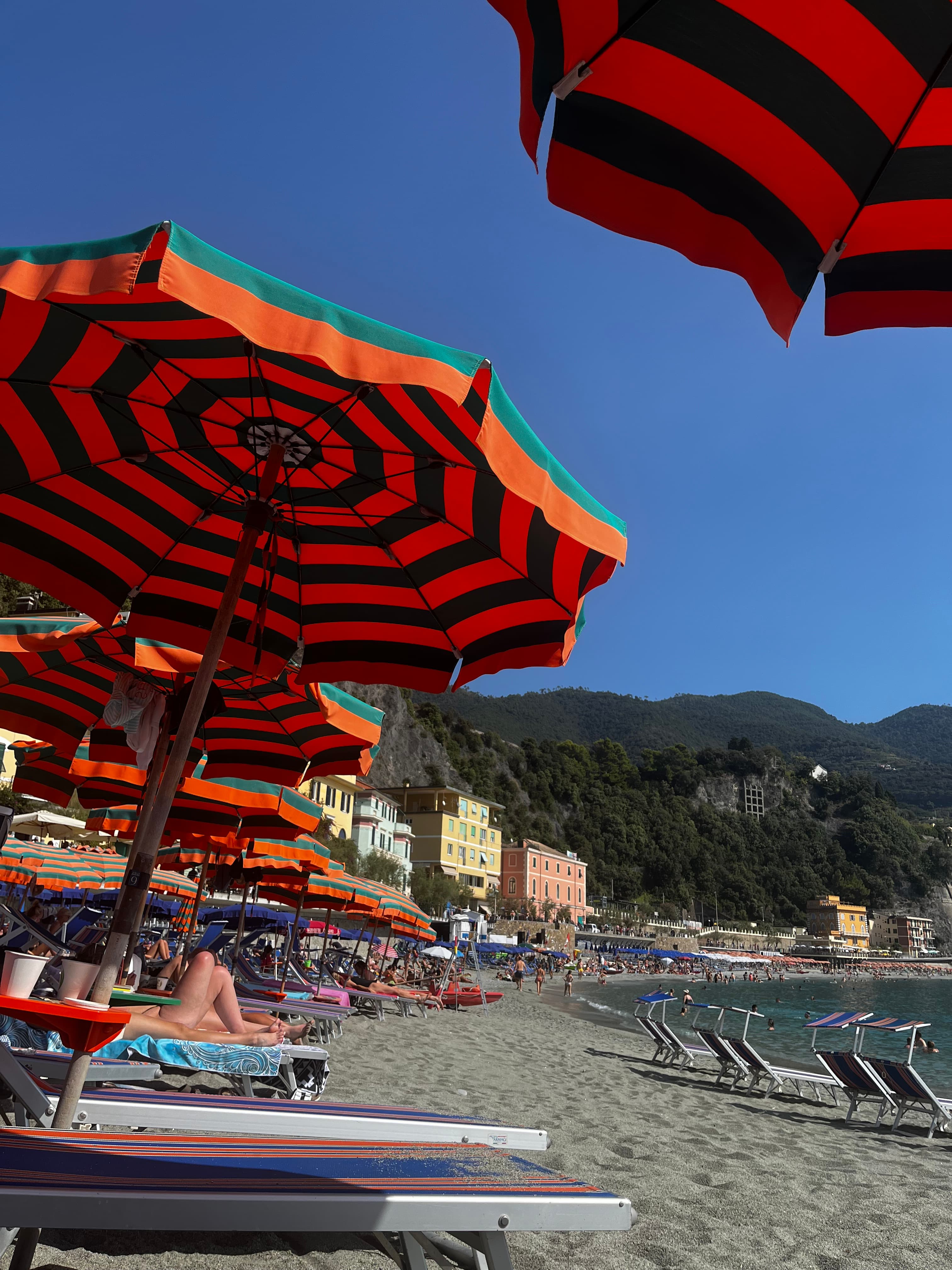 Red and black striped umbrellas and colorful lounge chairs on a beach with small buildings and green mountains in the distance.