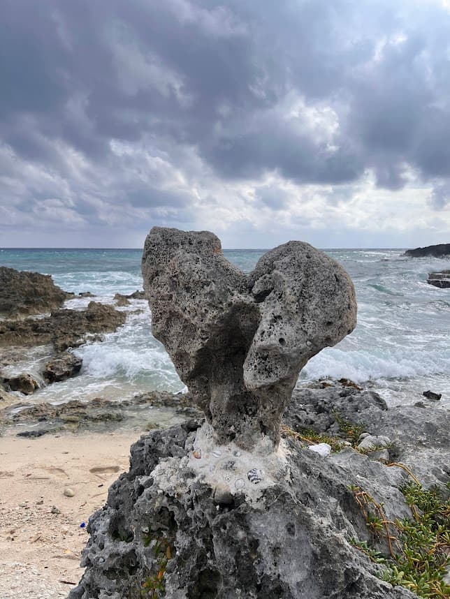An image of a hear shaped rock on the beach with sand and the ocean in the distance on a sunny day.