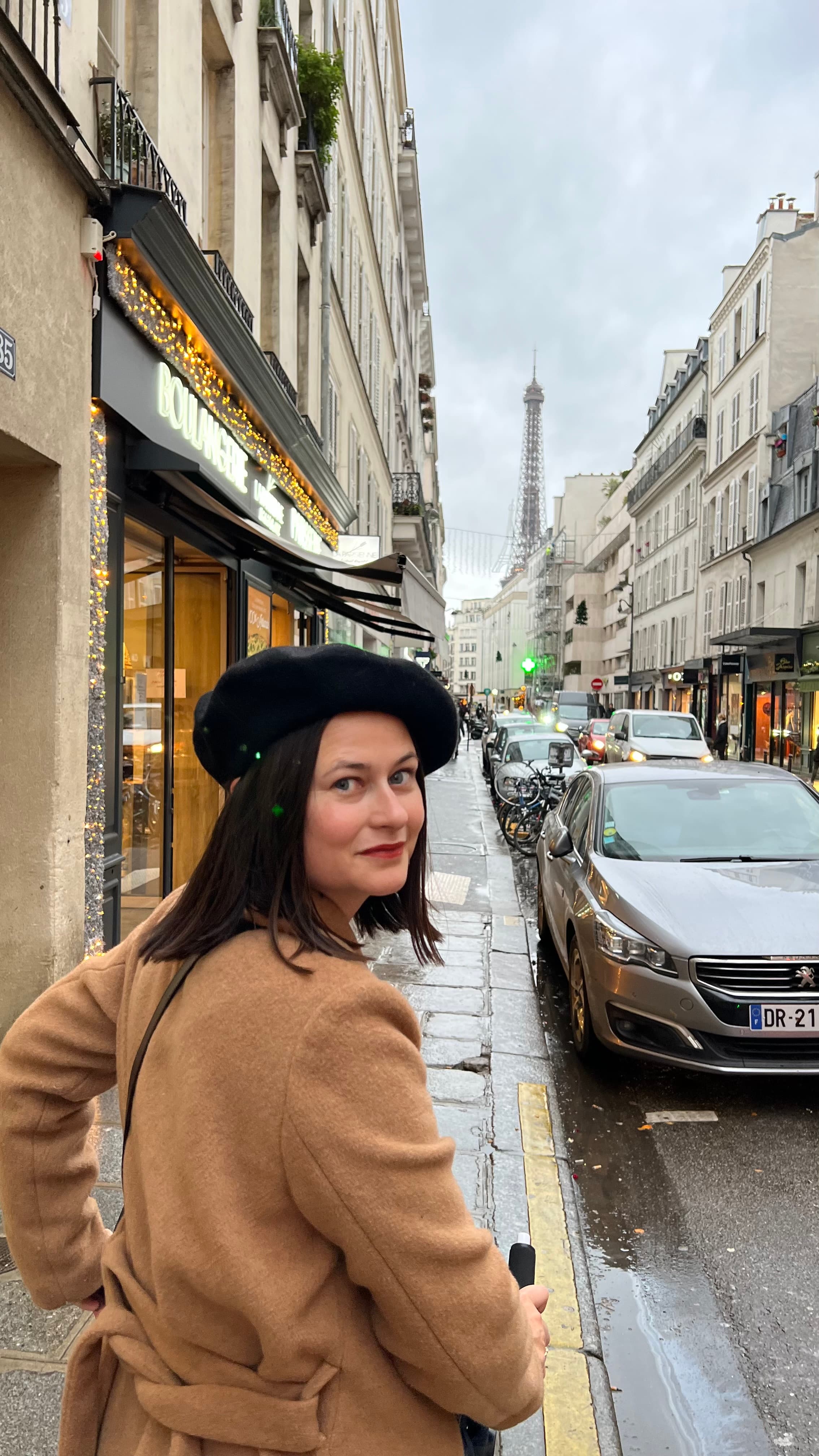 Photo of Samantha wearing a black beret and tan coat on the street in Paris with the Eiffel Tower visible in the distance
