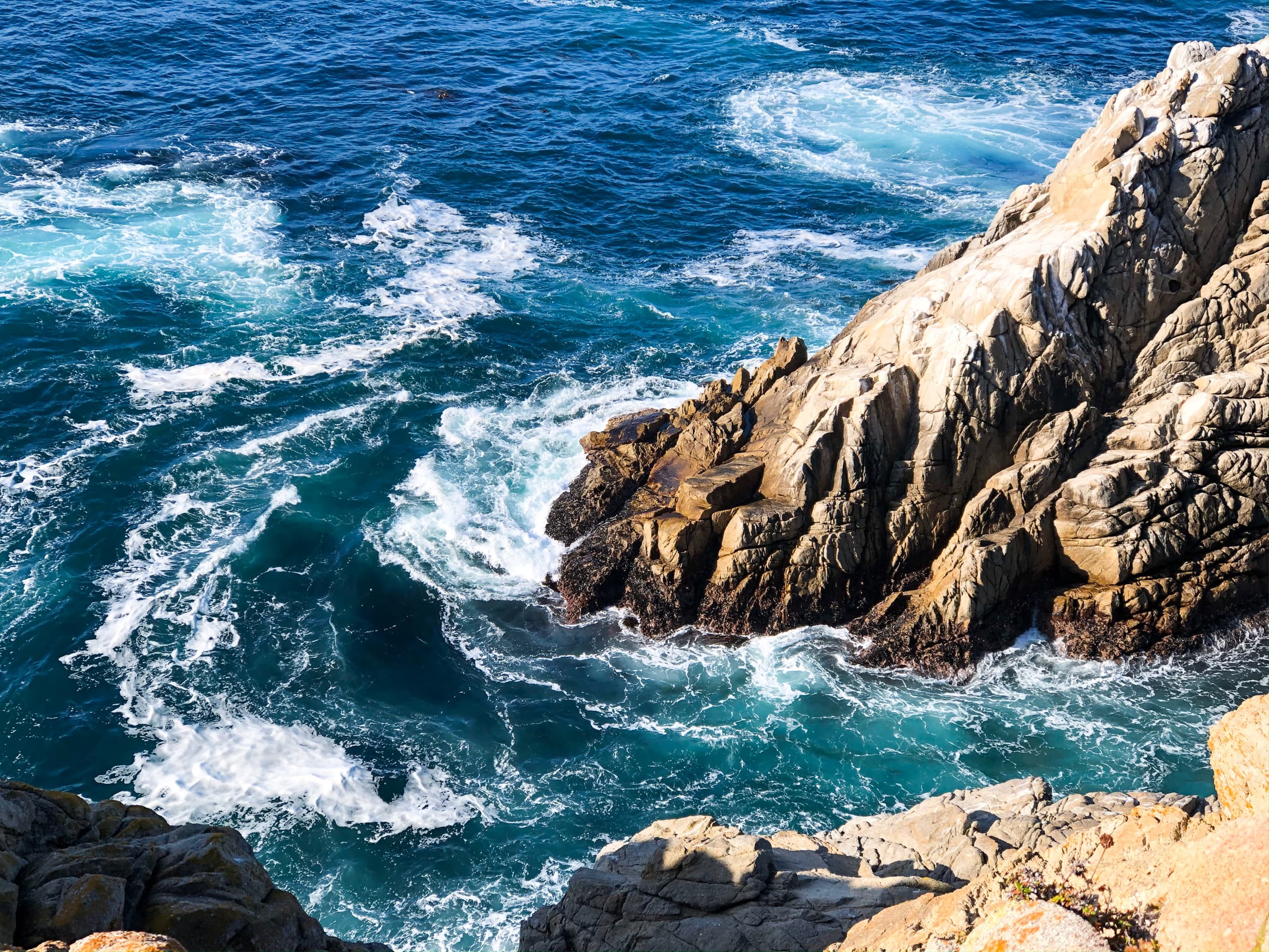 An aerial view of the ocean with rushing waves and descending cliffs into the ocean on a sunny day.