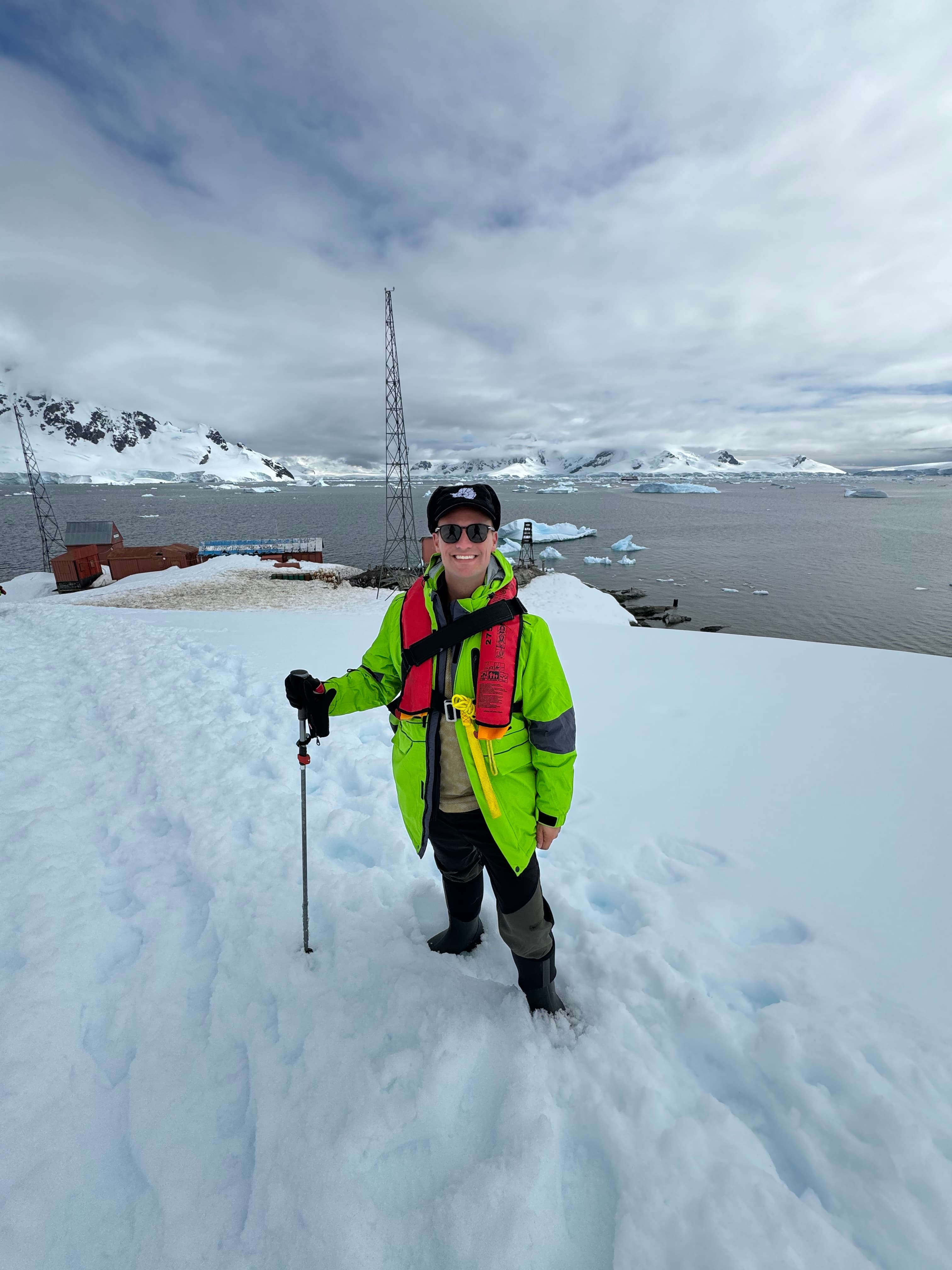 Alex in a bright green winter coat and black hat carrying a walking stick while standing on a snowy landscape