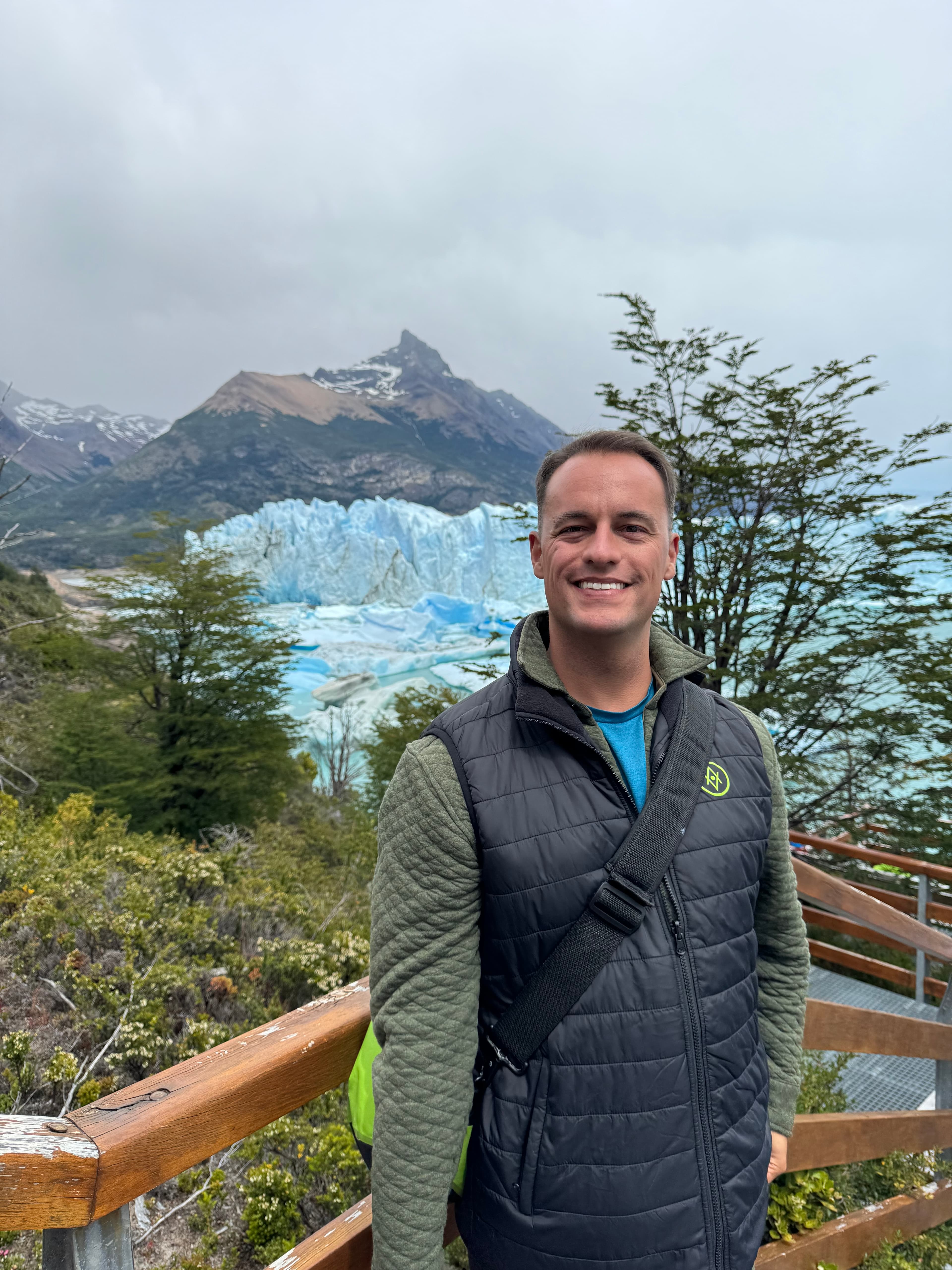 Alex wearing a vest and posing on a wooden platform with a beautiful view of glaciers and mountains behind him