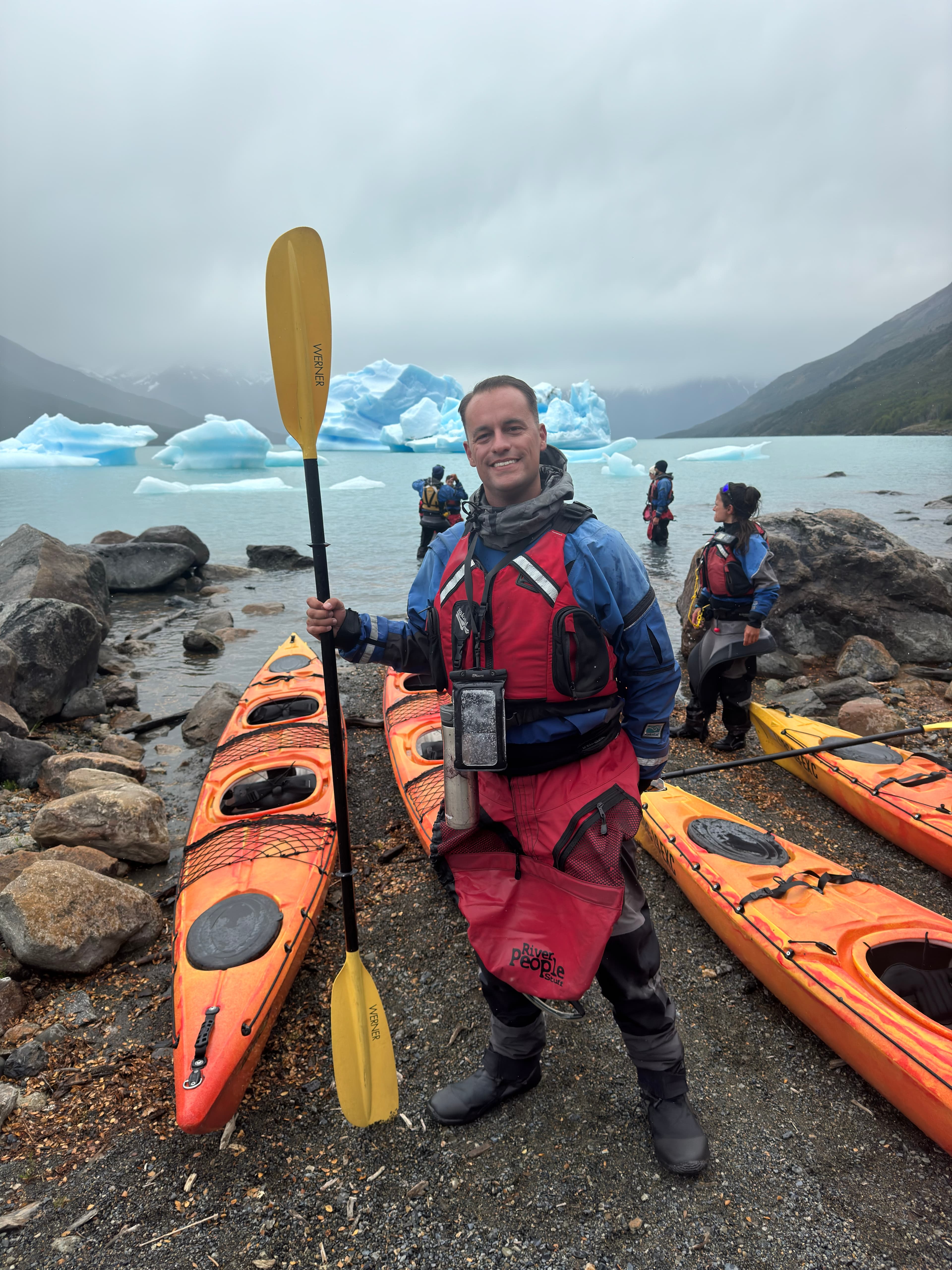 Alex in winter clothing holding a yellow oar by several orange kayaks with large glaciers visible behind him
