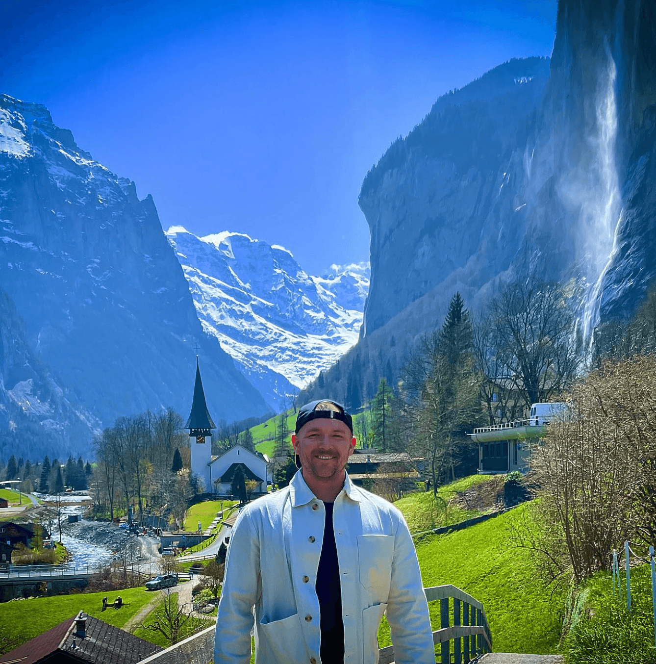 Andrew in a white jacket posing in front of a beautiful alpine landscape with snowy mountains on a sunny day