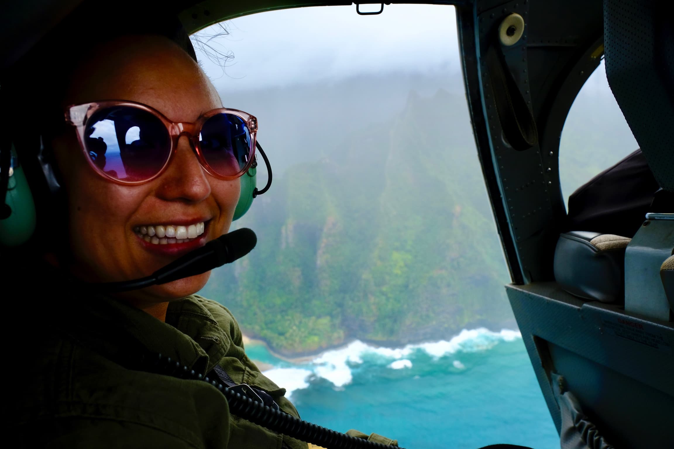 A person smiling with a pilots headset on, flying over a body of water. Waves crashing on the beach and forest in the distance.