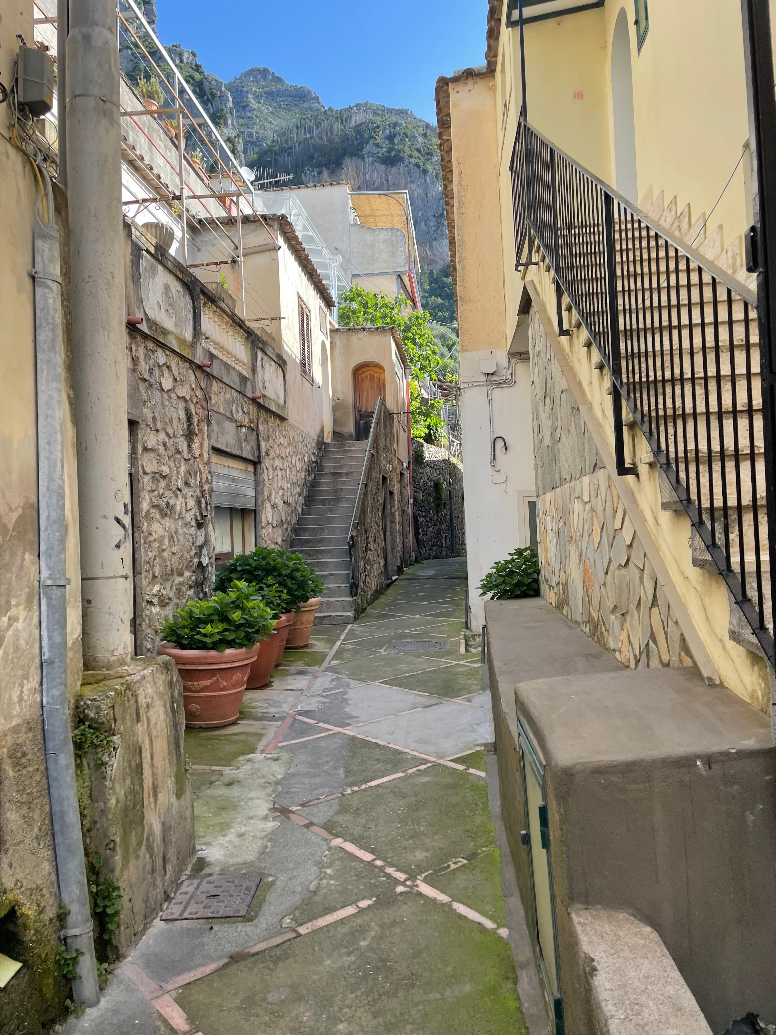 View of a narrow stone pathway leading in between small buildings with staircases on either side on a sunny day