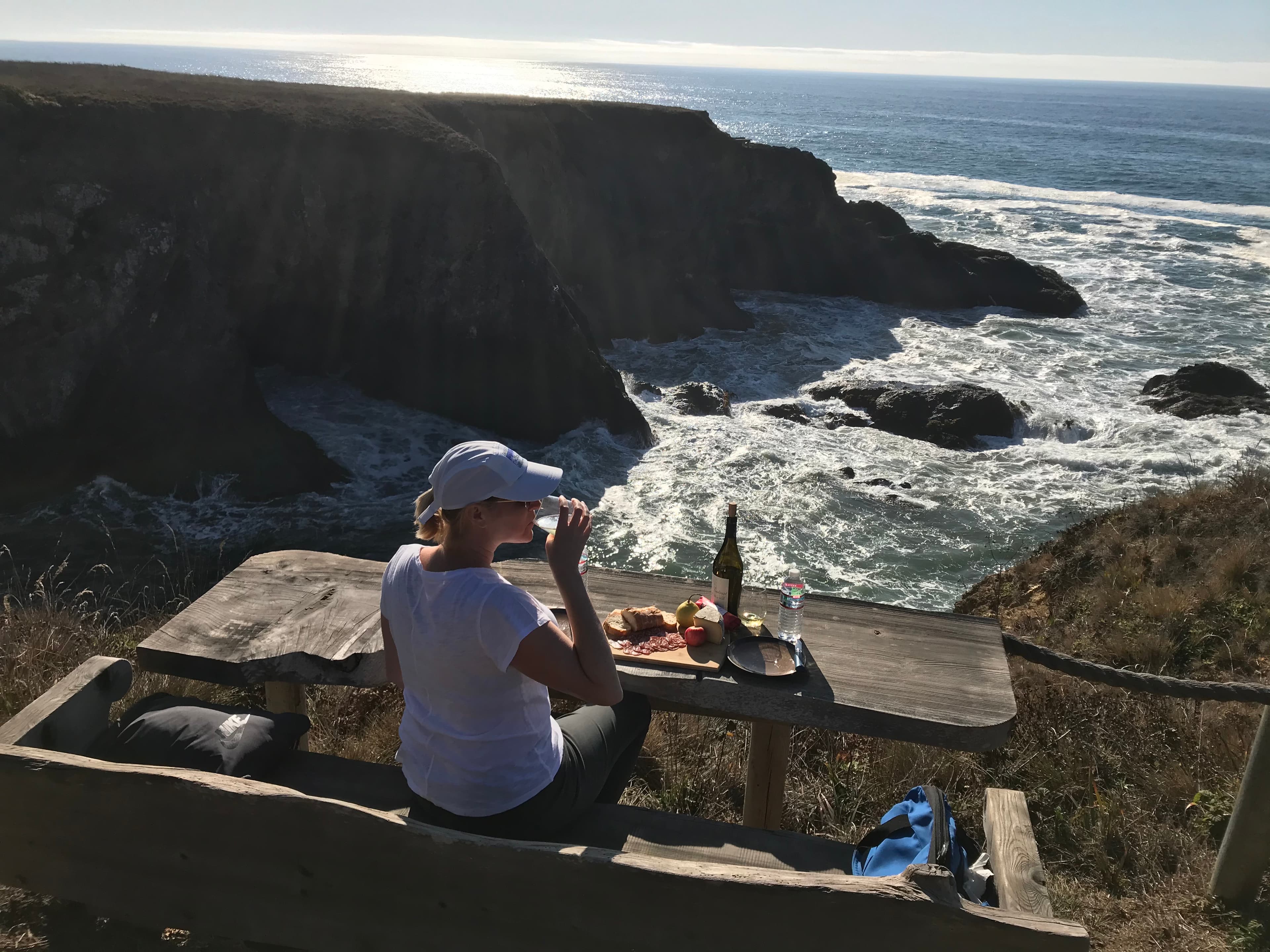 Leslie seated at a lookout point with food and a bottle of wine overlooking a beautiful ocean view