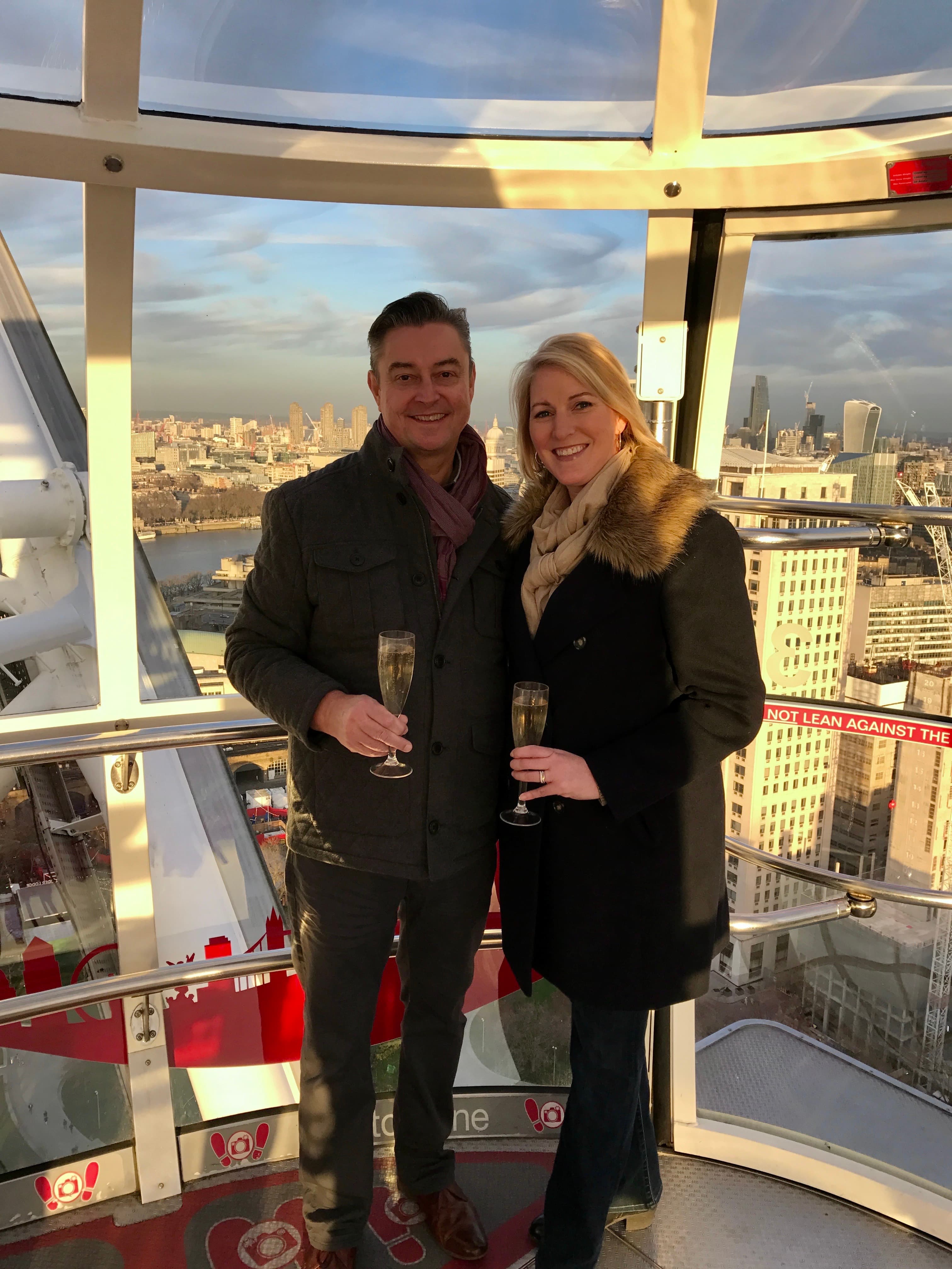 Leslie and her husband holding champagne flutes and posing in front of glass windows, a cityscape visible behind them