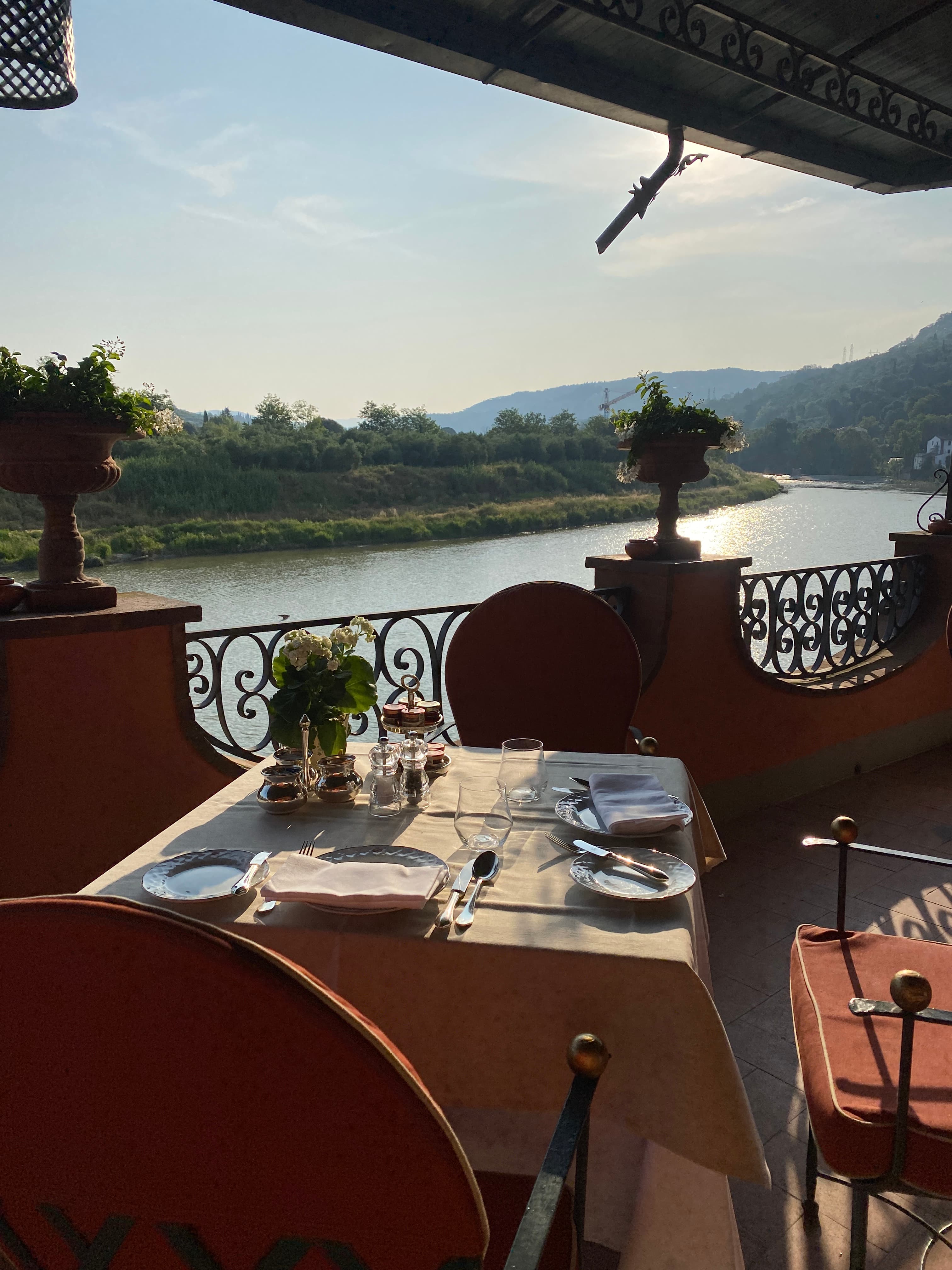 An outdoor restaurant table set for two on a balcony overlooking a beautiful river on a sunny day
