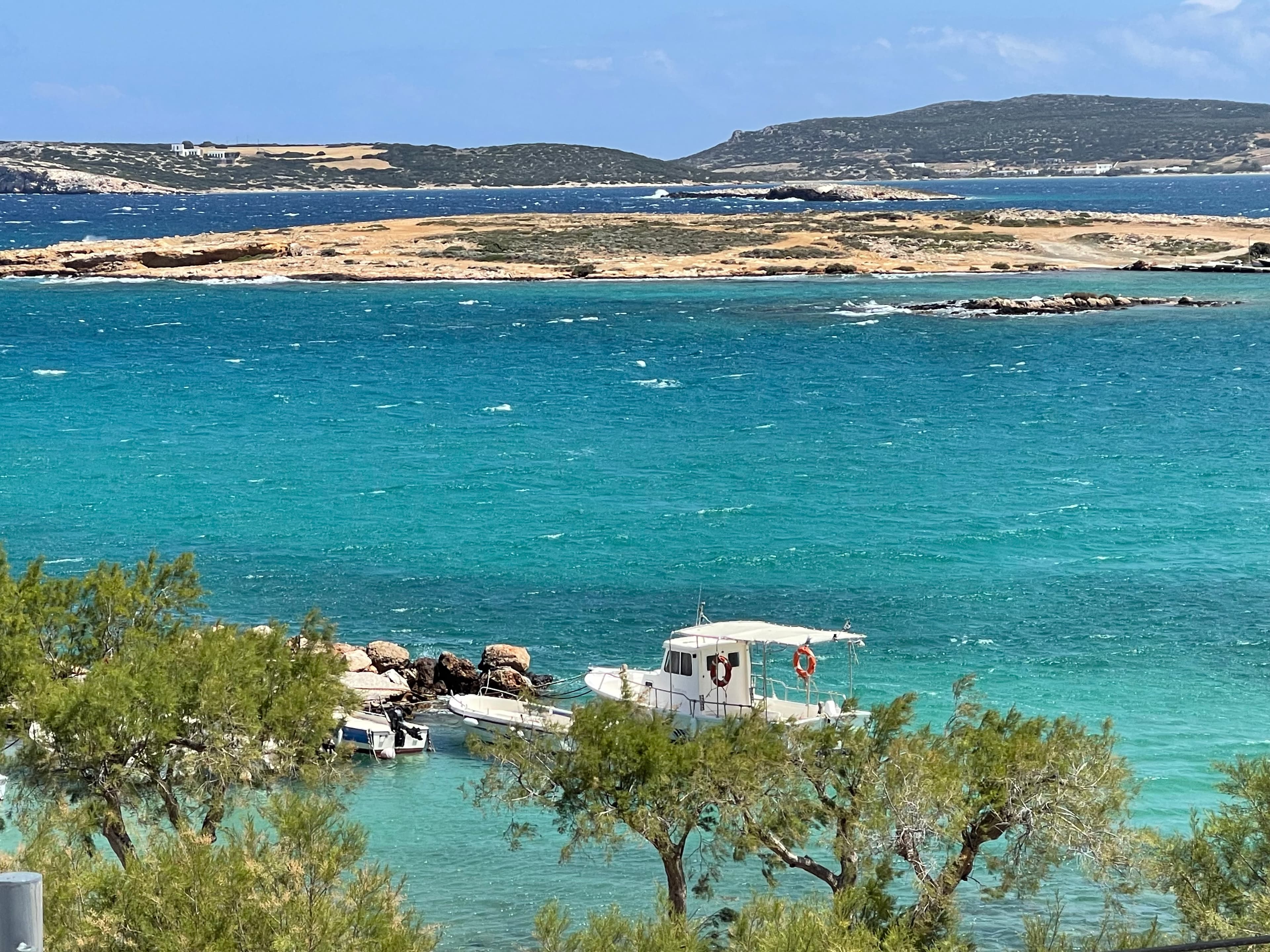 Small white boat anchored in turquoise water near a rocky shoreline with trees in the foreground and distant islands under a blue sky.