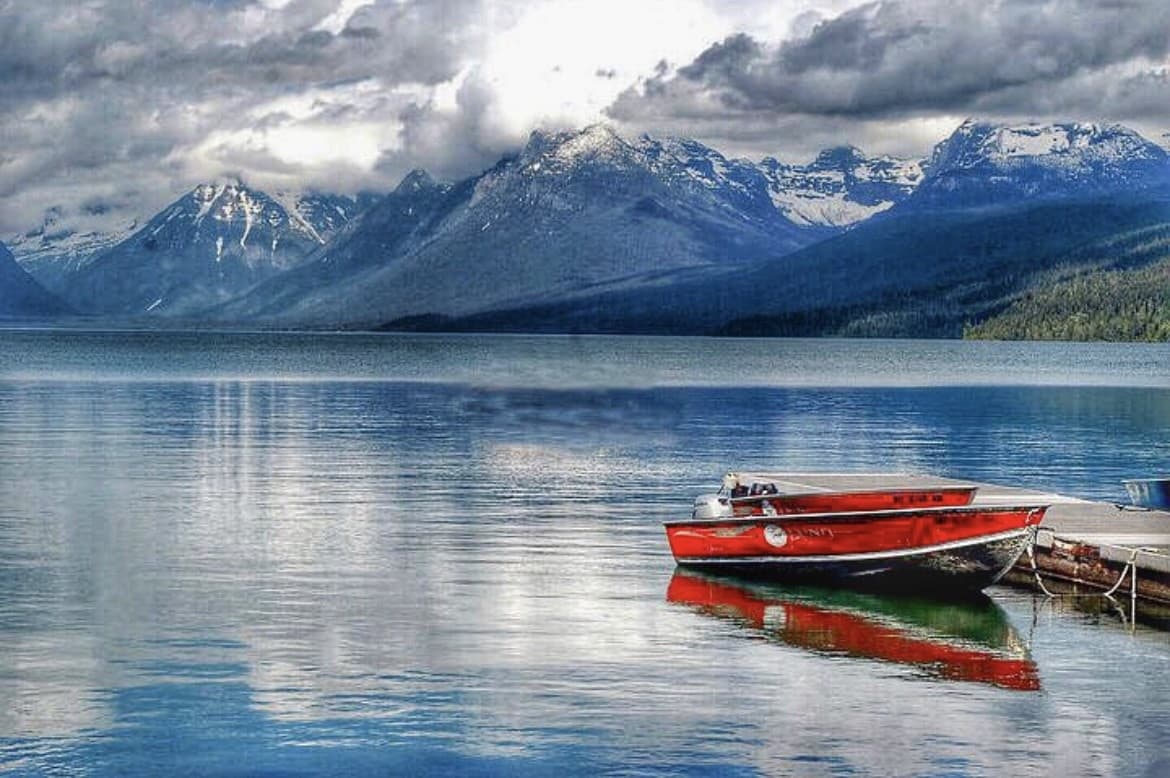 A boat rests on still waters as clouds cascade down a mountainside on the shore.