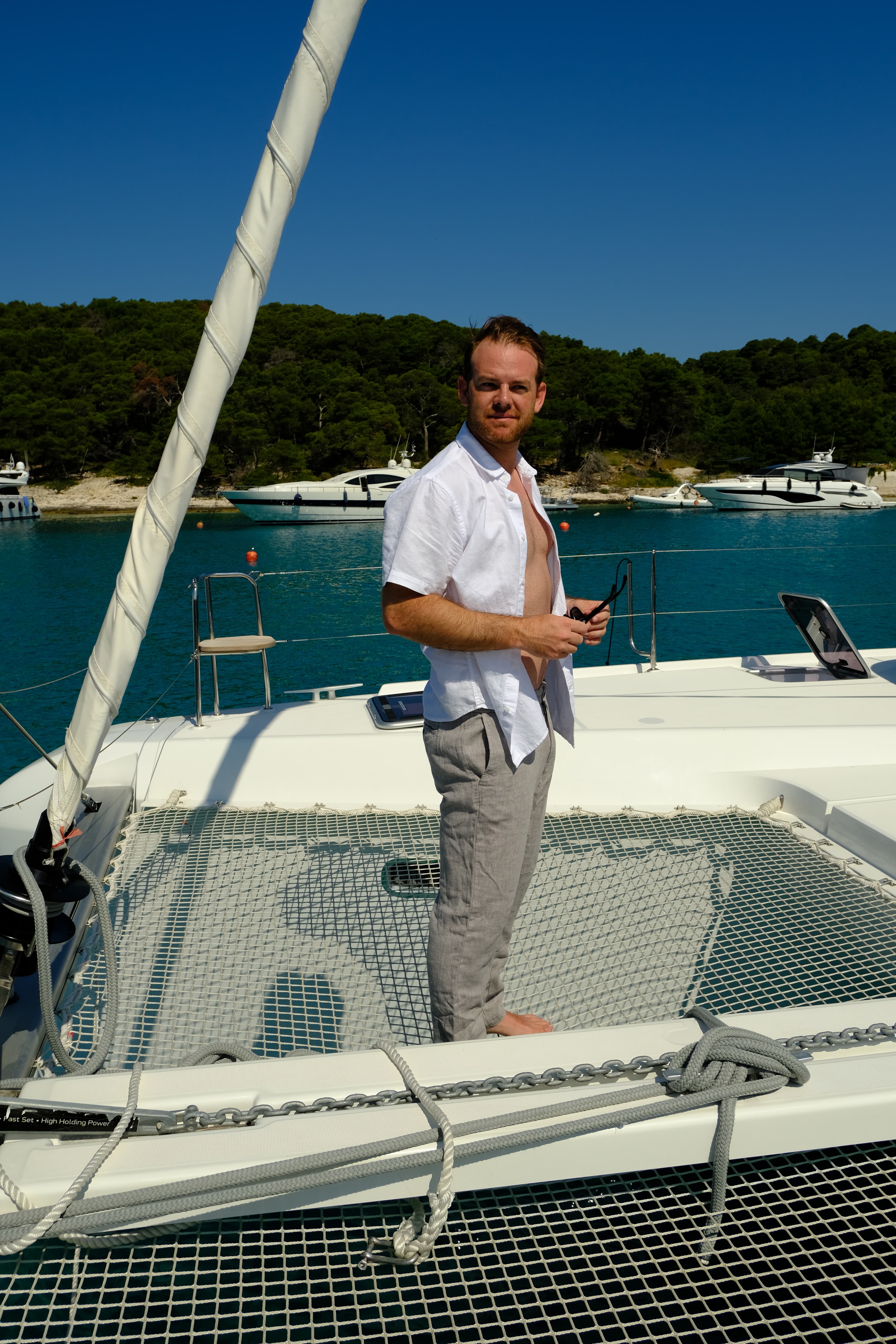 A man posing on a boat while wearing a white shirt unbuttoned and grey pants.