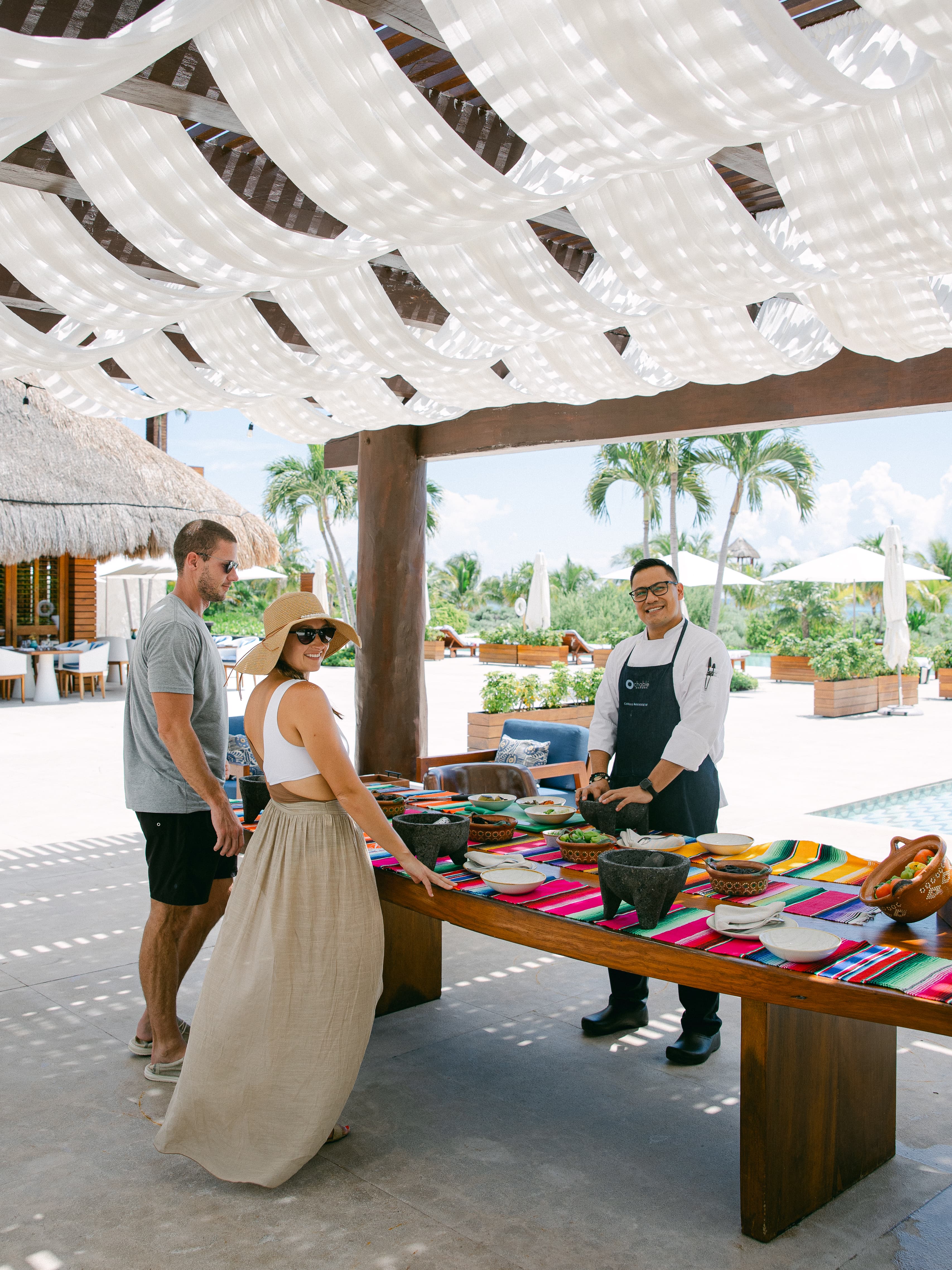 A couple posing outside in front of a table where someone is selling goods. There is a white sun covering above them.