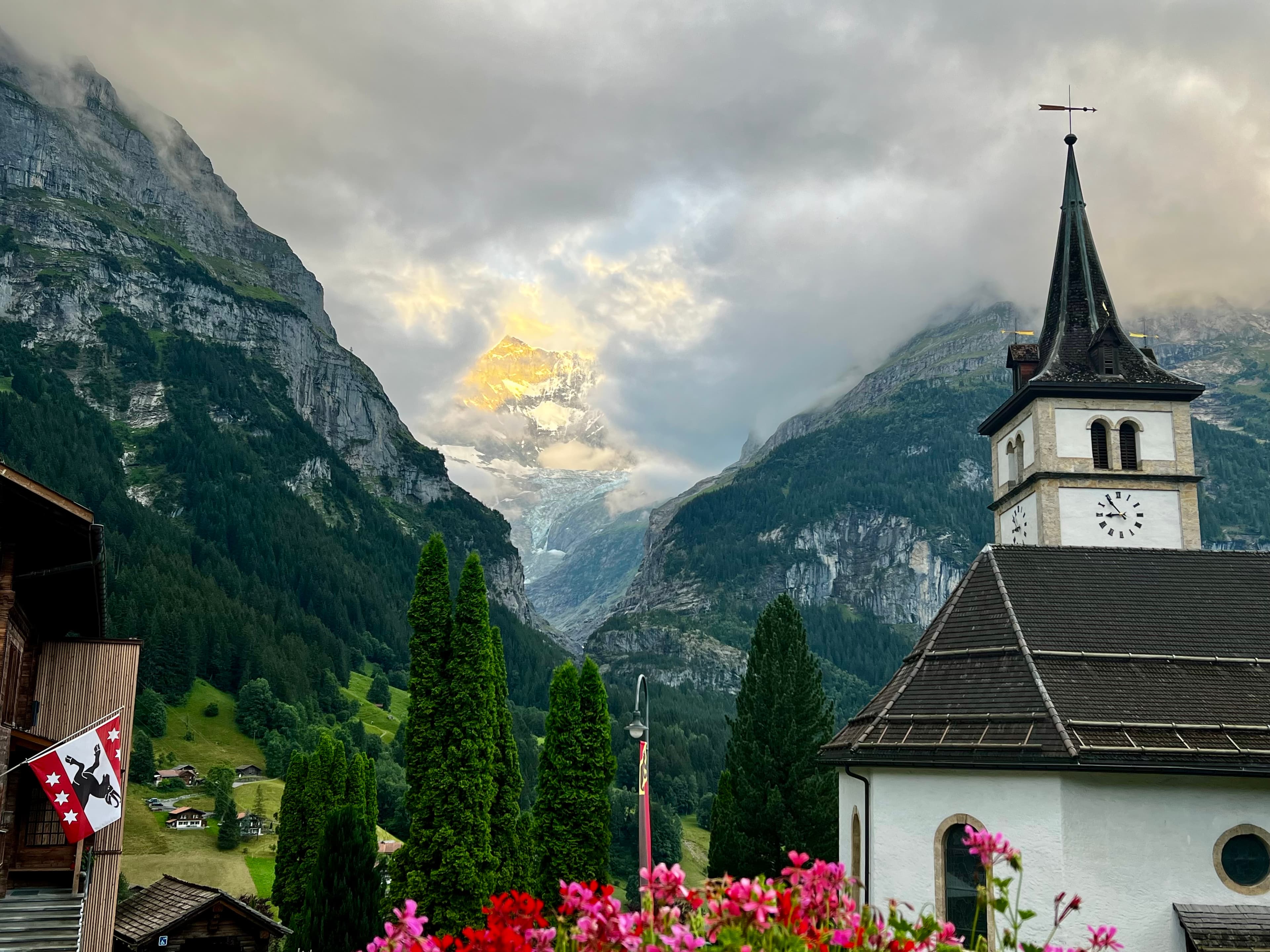 A church featuring a prominent steeple, set against a picturesque mountain backdrop under a clear blue sky.