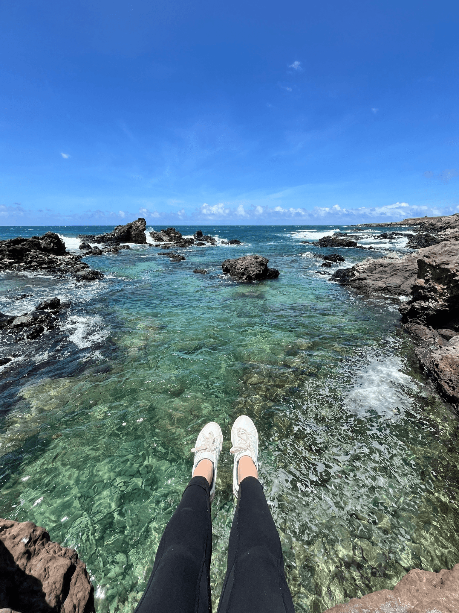 View of advisor’s legs dangling off a cliff overlooking clear ocean water on a sunny day