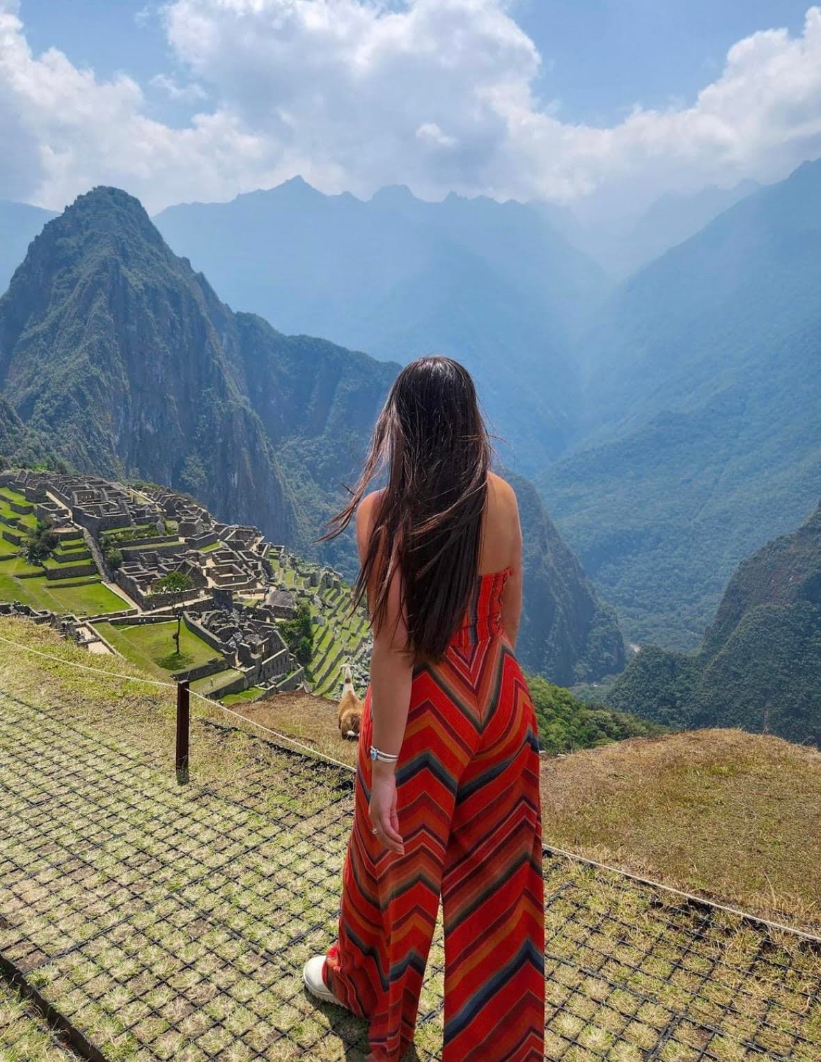 Caity wearing an orange dress stands at the terrace's edge, admiring the stunning ruins of Machu Picchu in the distance.