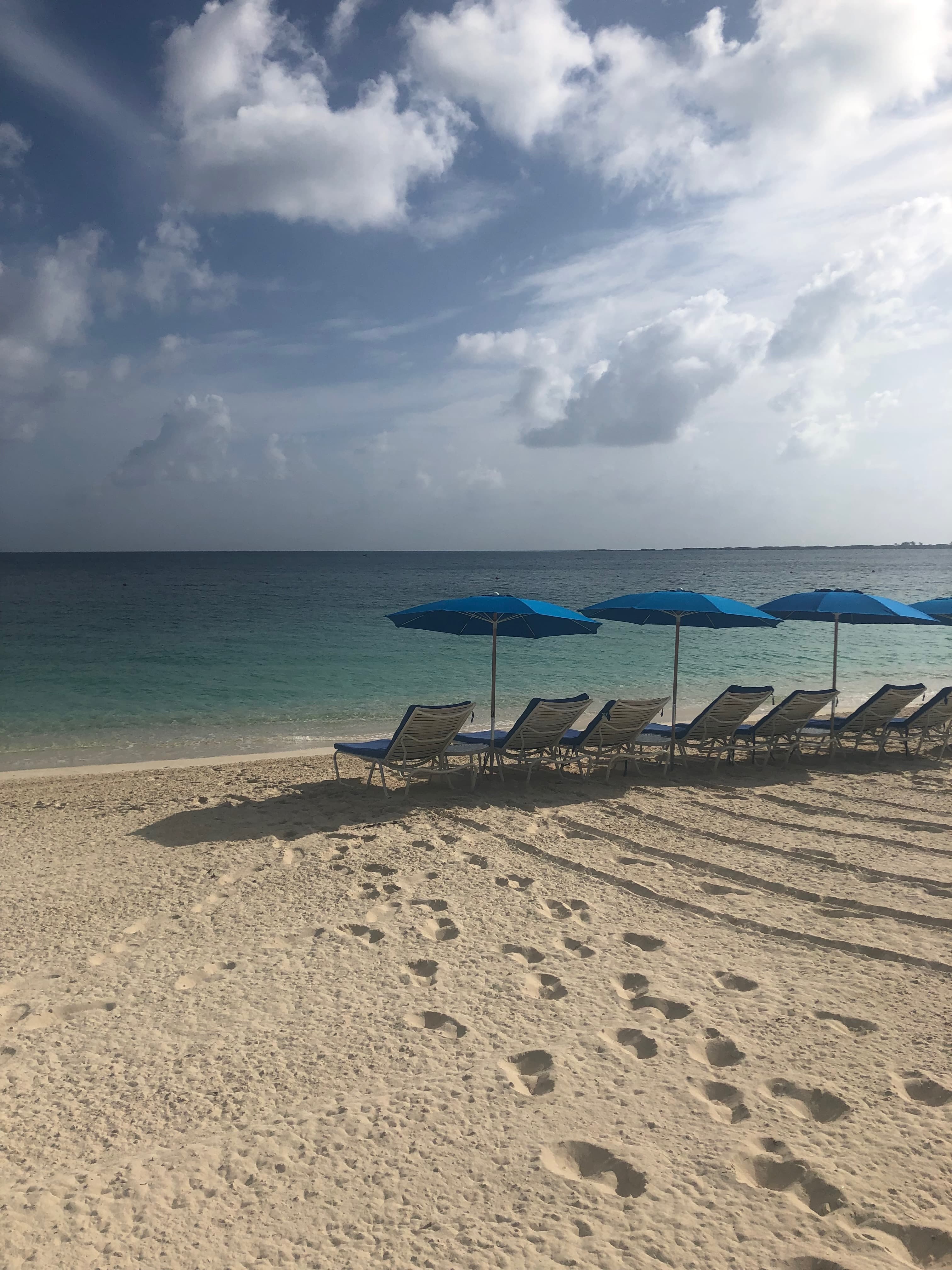 View of footsteps in the sand of a beach and a line of empty lounge chairs by the water’s edge