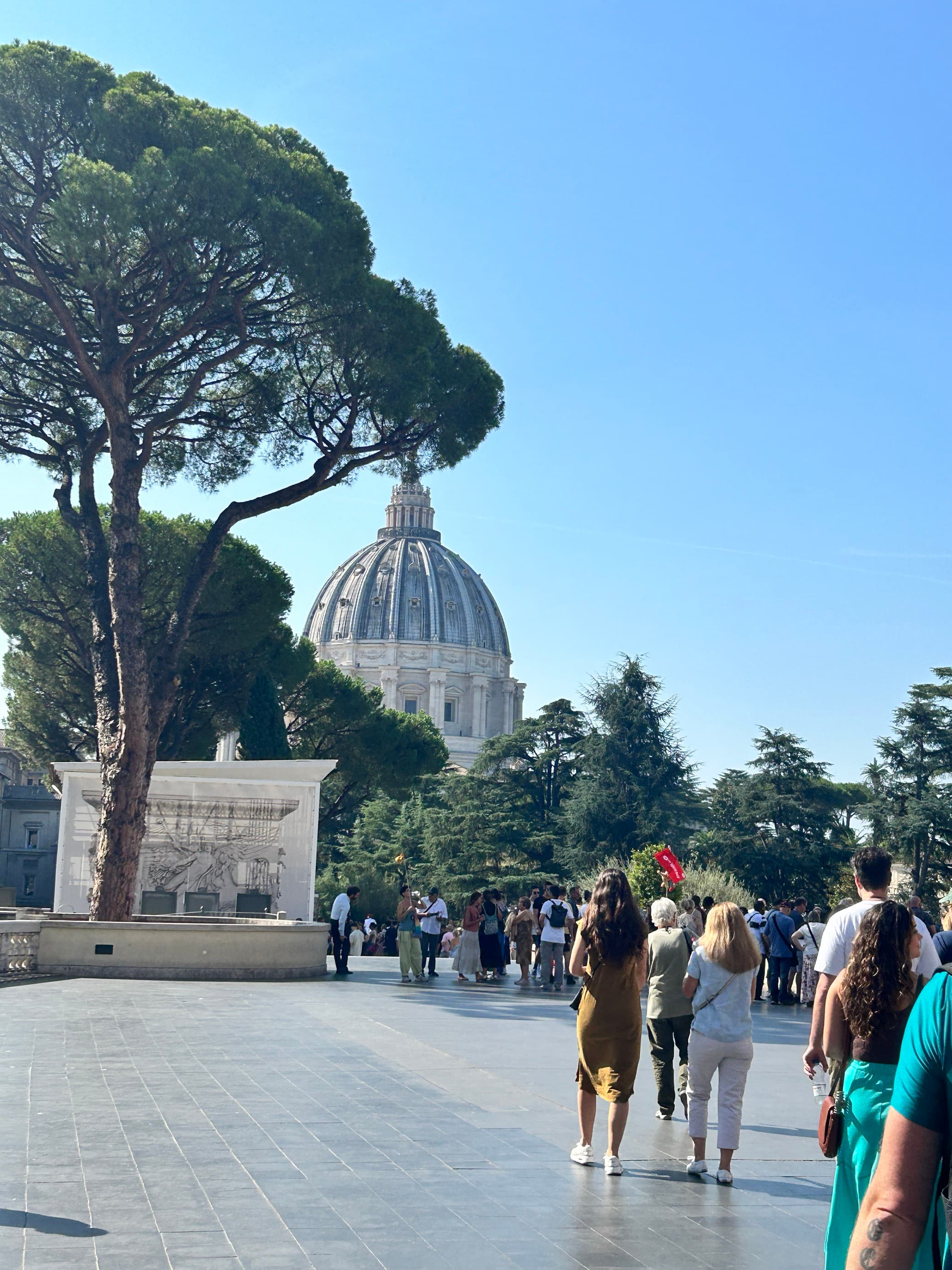 View of pedestrians walking through a sunny city plaza with trees and a large domed building in the distance