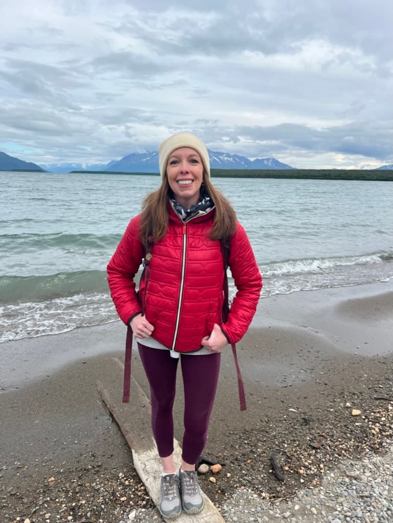 Advisor in a red jacket and white winter hat on a beach with the sea and mountains in the distance