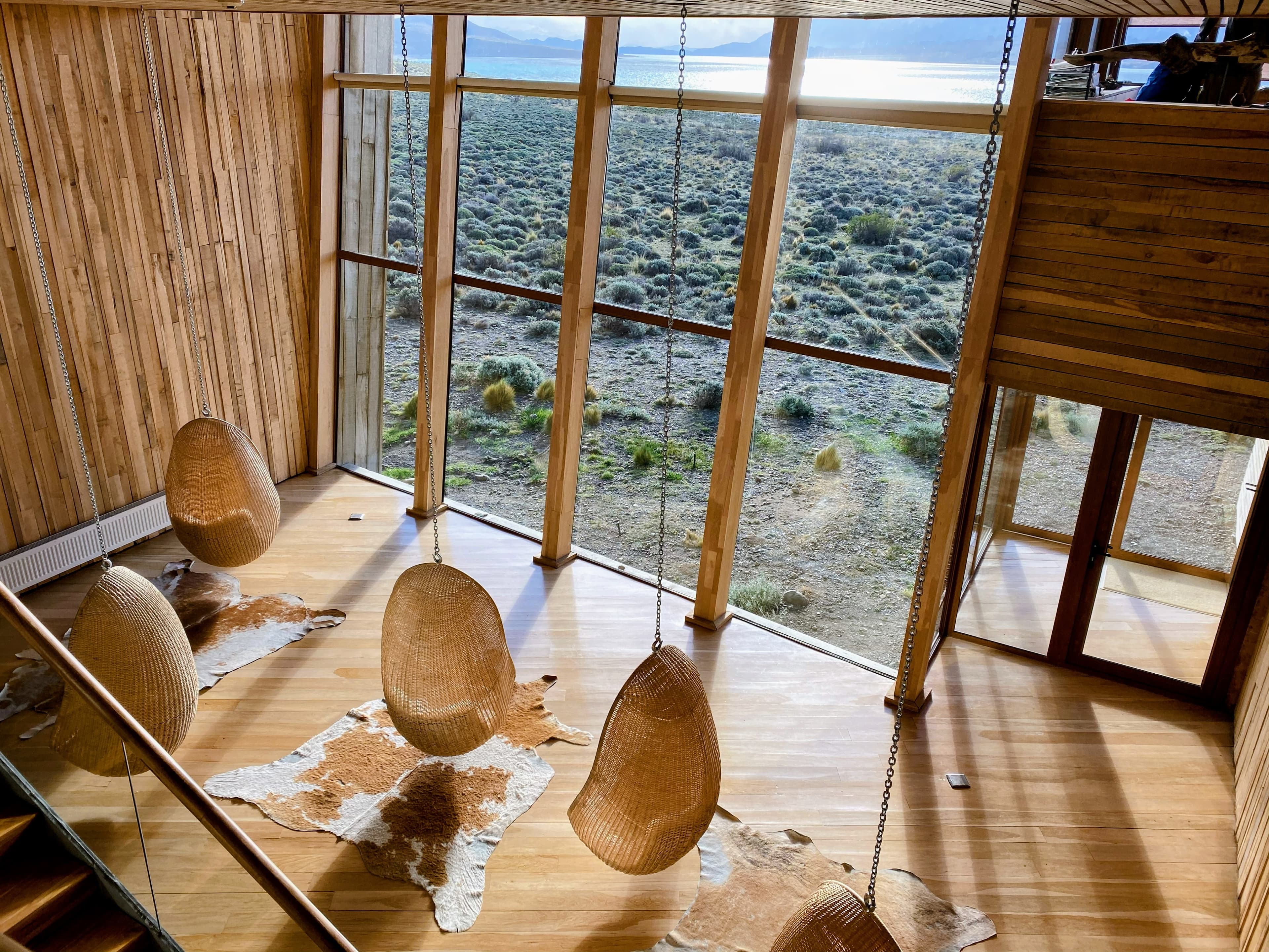 View of hanging chairs in a wooden room with floor to ceiling glass windows overlooking a grassy area