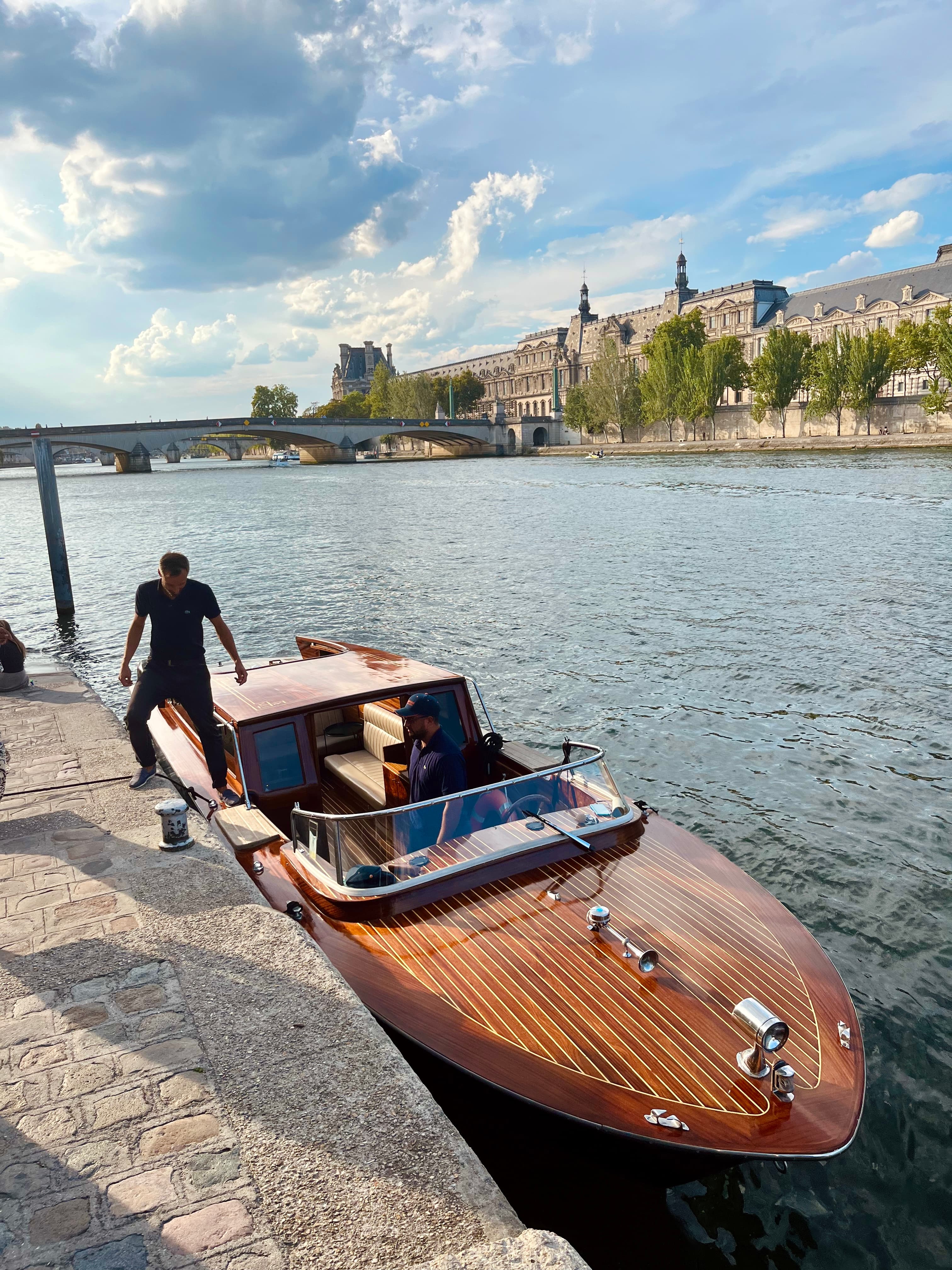 View of a small brown boat docked along the side of a river pier with a person stepping out on a sunny day