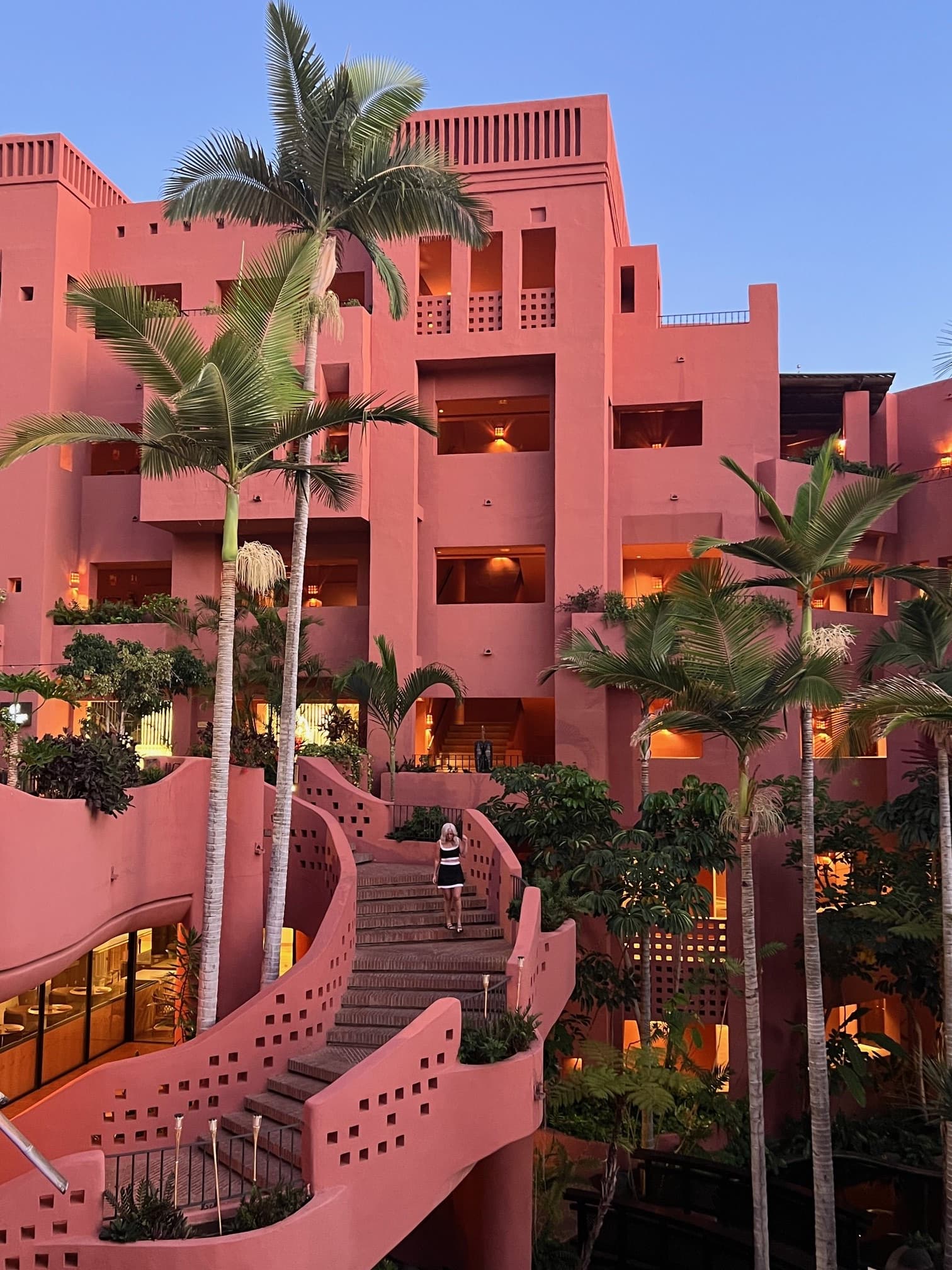 View of a pink resort building with a curved staircase and nearby palm trees on a clear evening