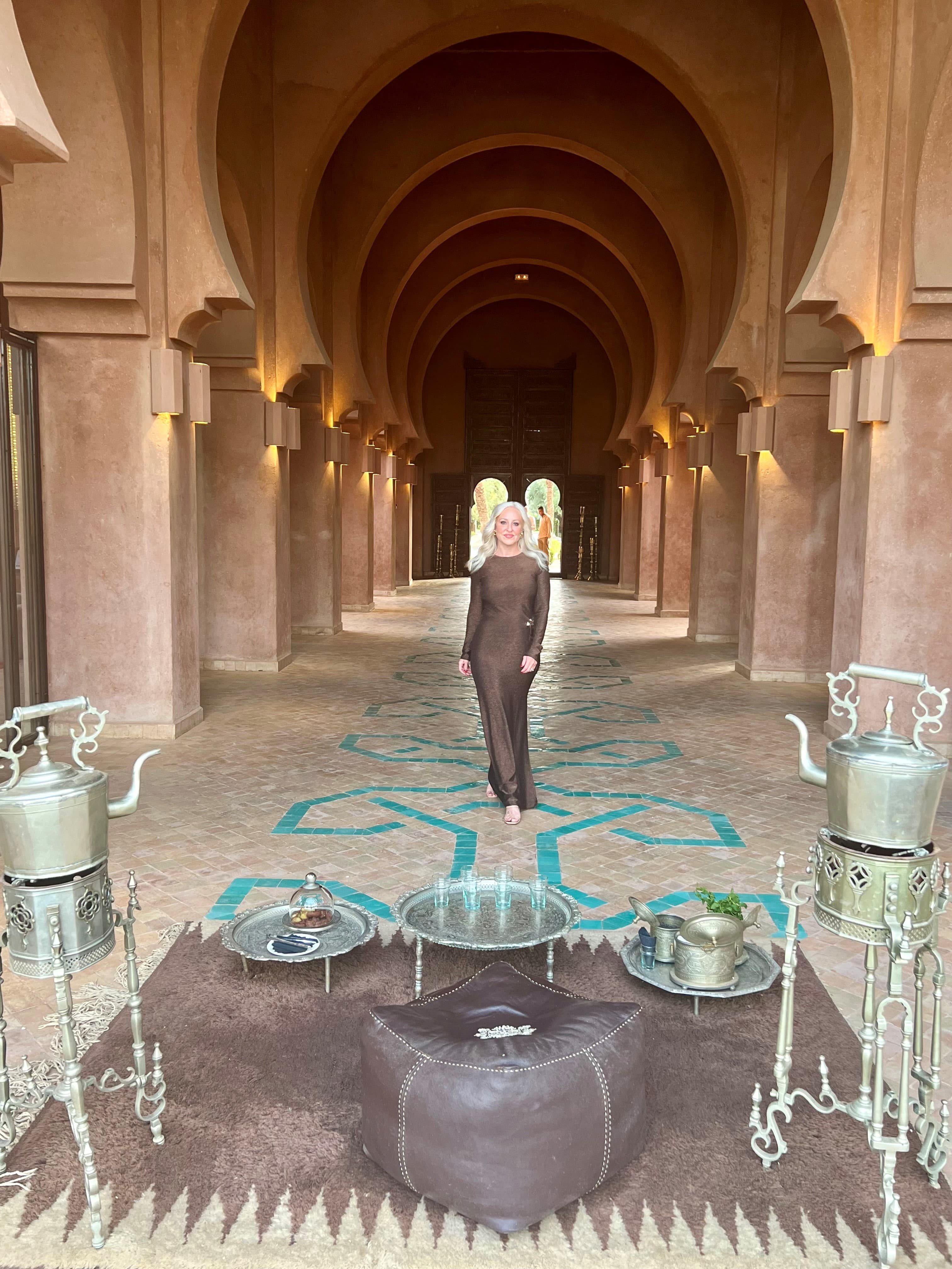 Advisor standing in the center of a grand hall lined with square columns with platters of tea on a carpet behind her