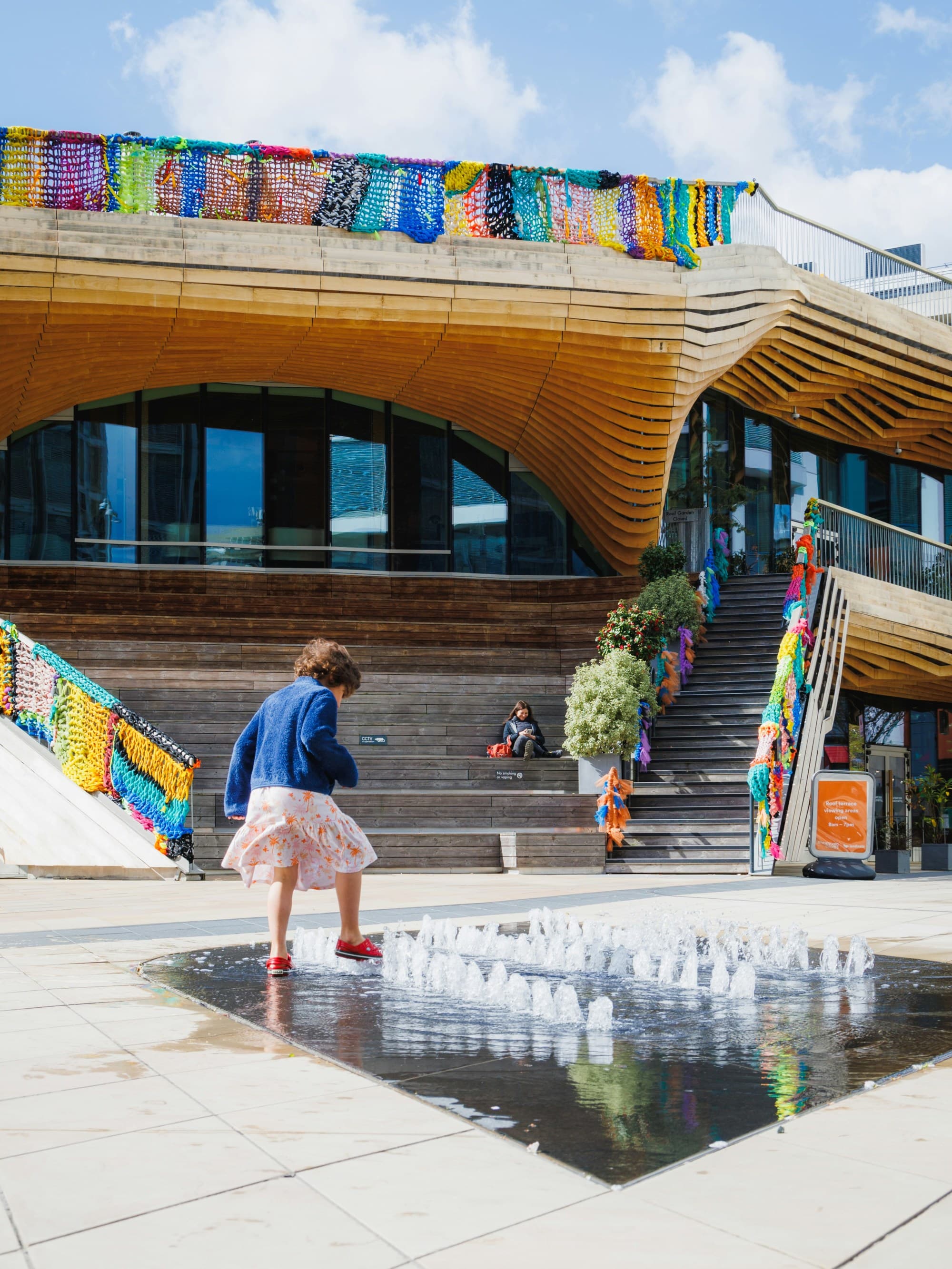 A modern building with a wooden facade and colorful banners, featuring a fountain and people sitting on steps.