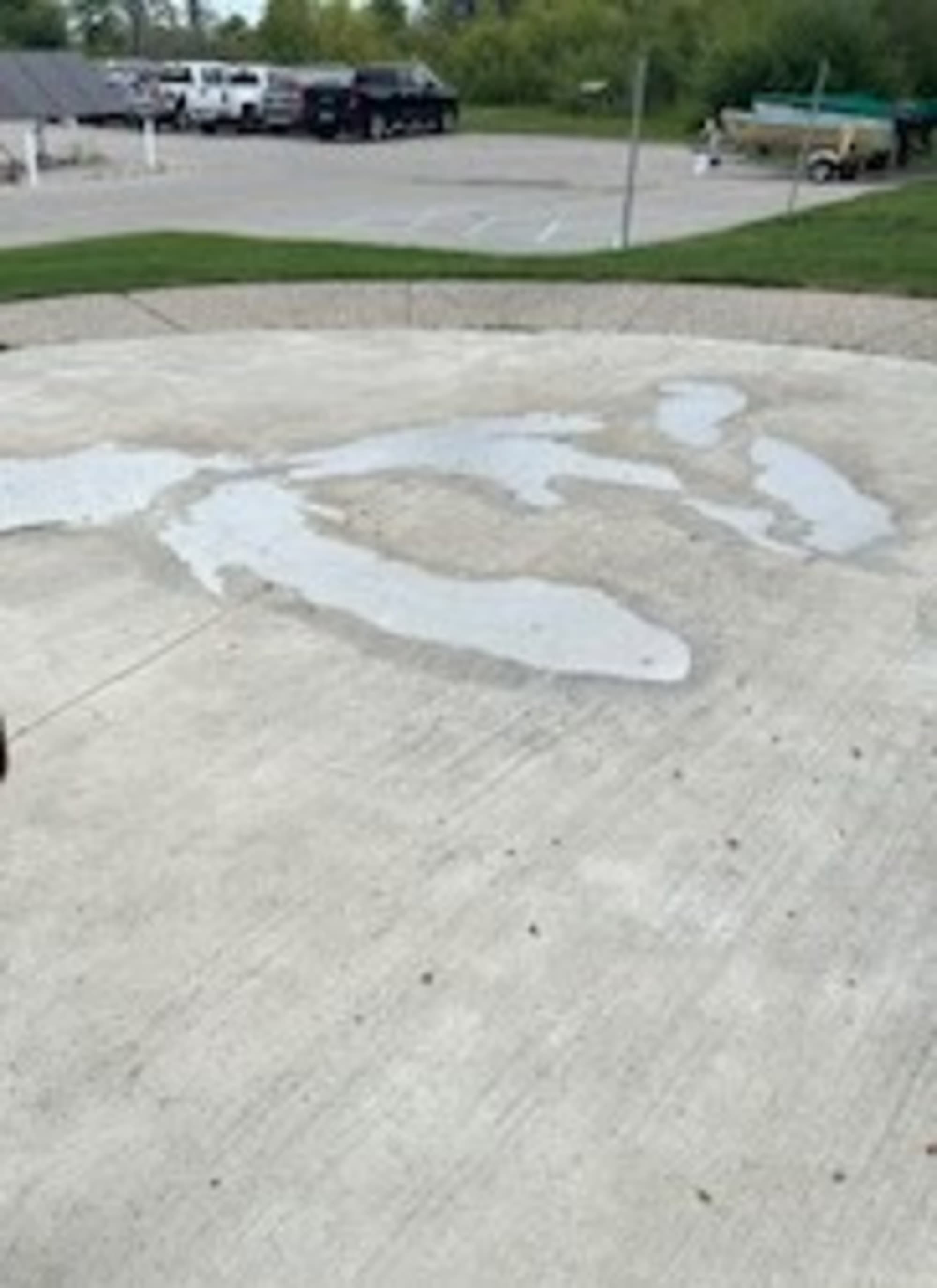 A concrete surface with irregular wet patches, vehicles, and greenery in the background.