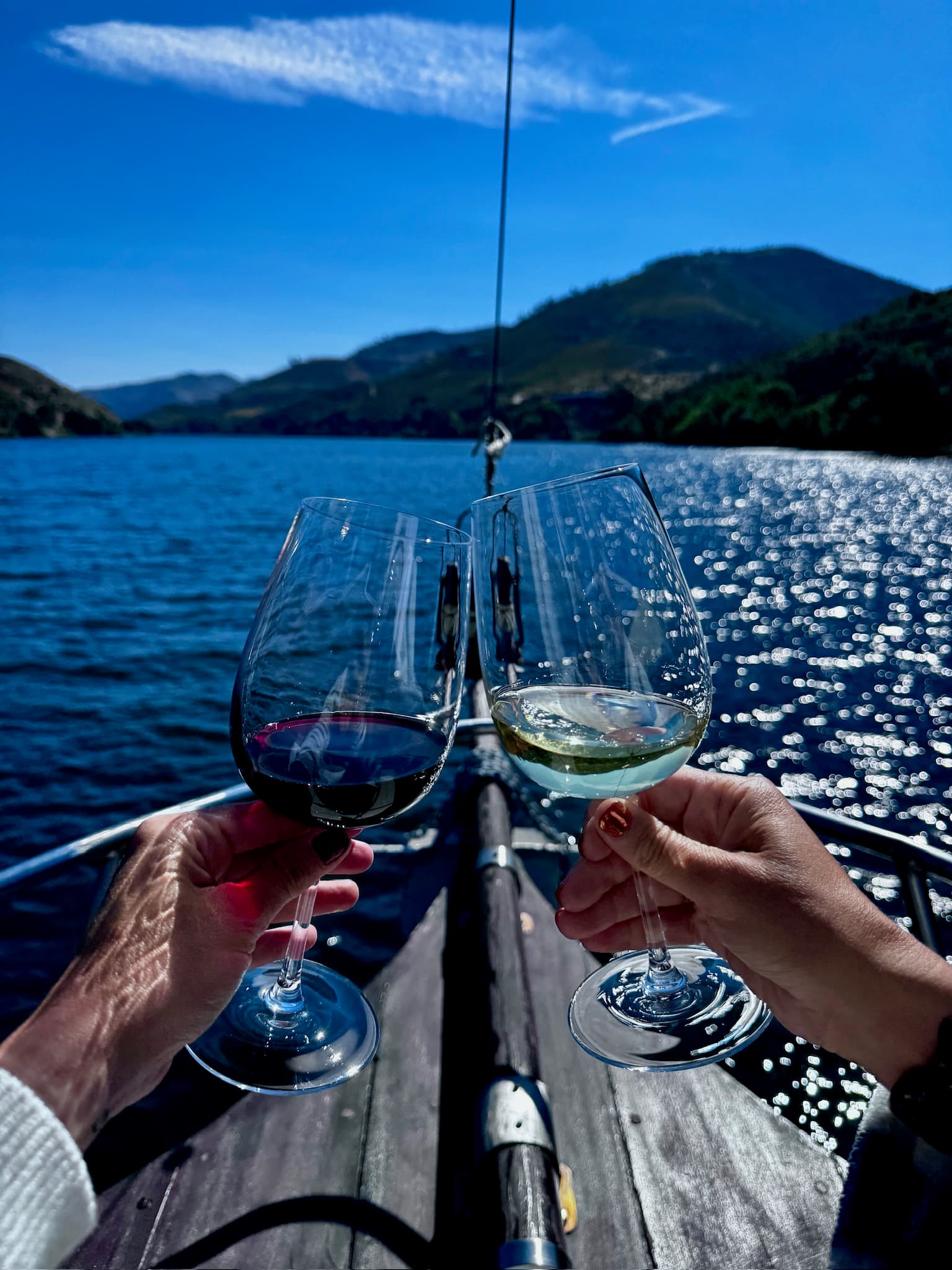 Two people's hands clinking wine glasses while on a boat in a river during the daytime