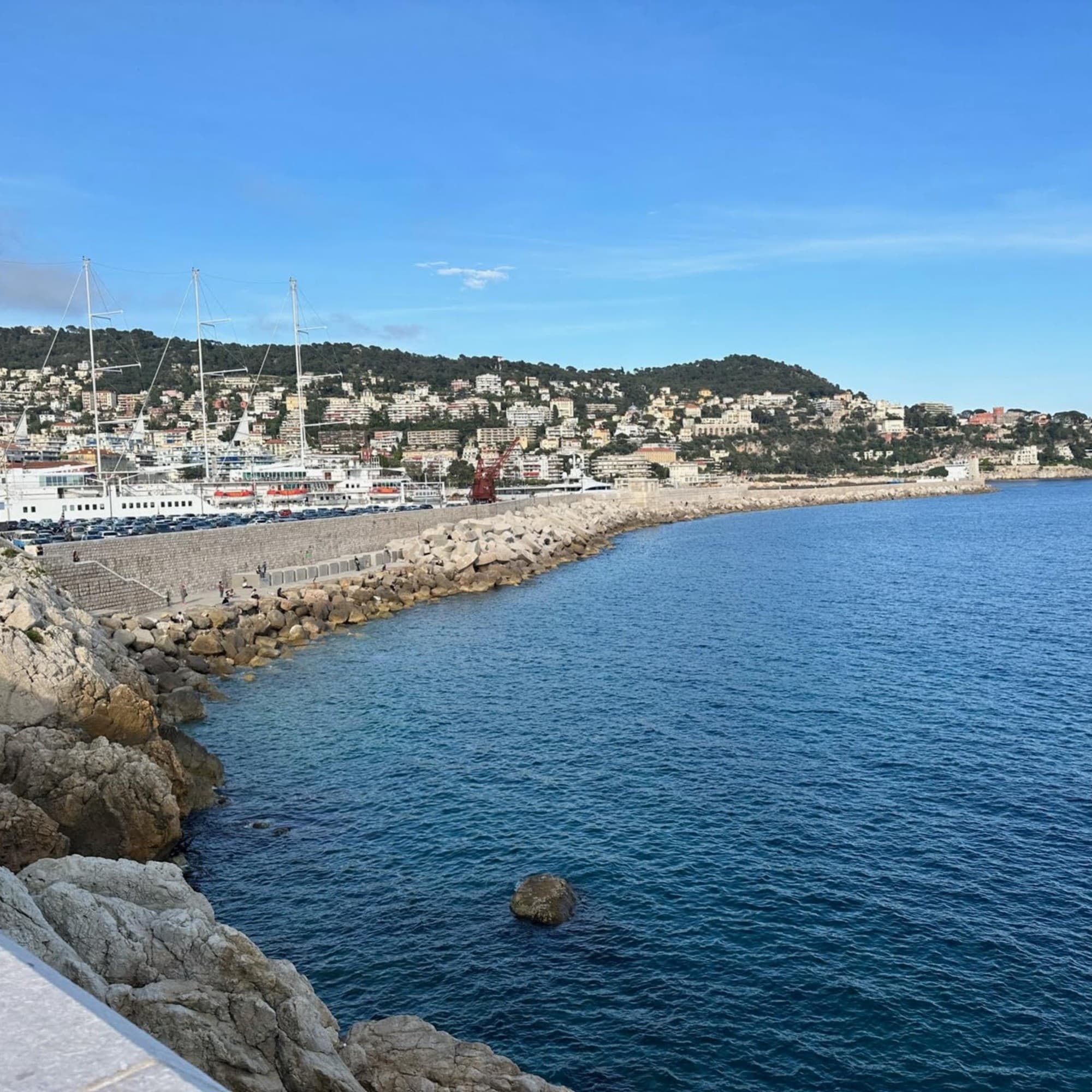 A coastal seascape with calm blue water, a cityscape along the shoreline and boats under a clear blue sky.