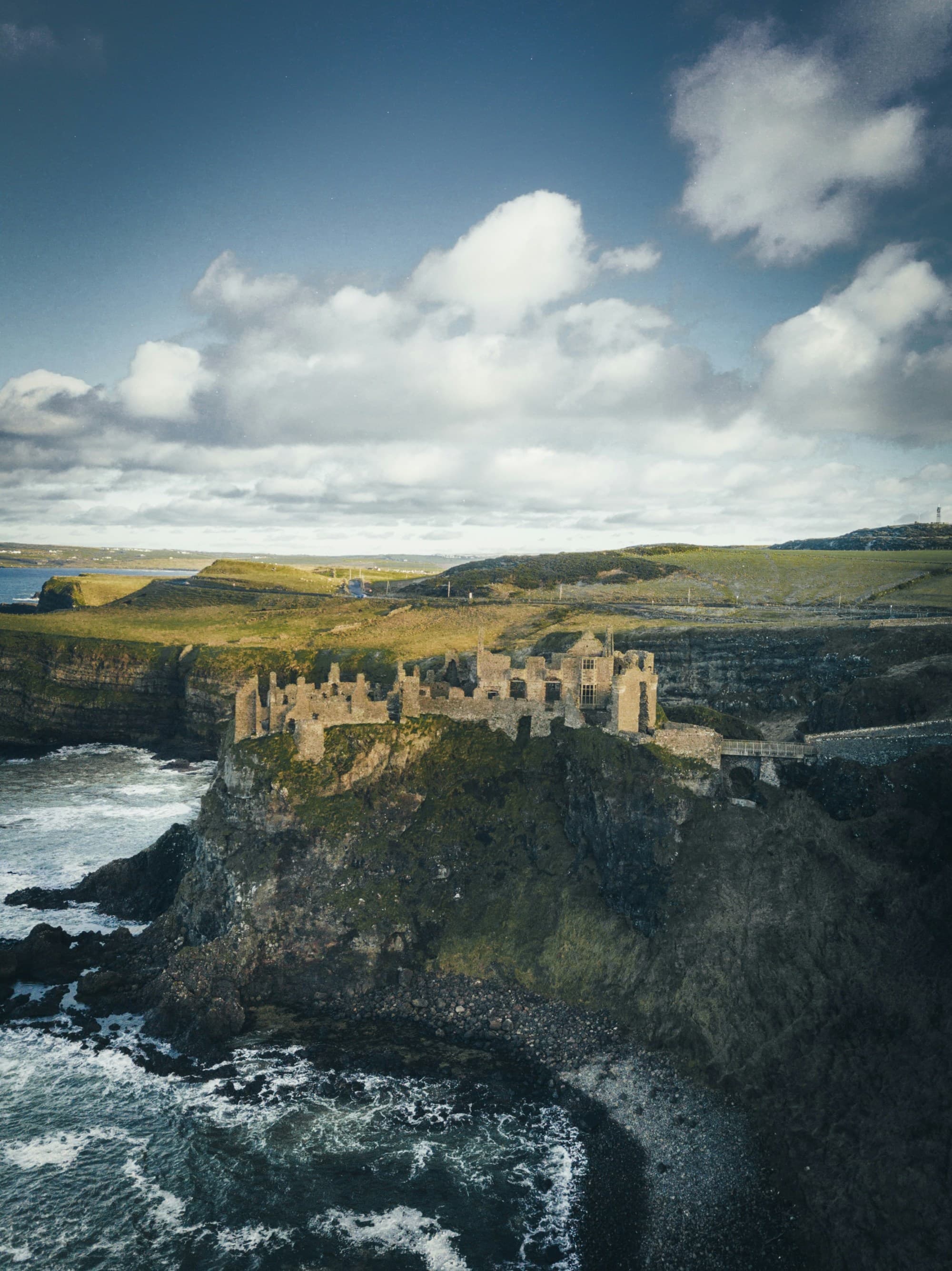 An aerial view of a ruined castle on a rugged cliff with ocean waves crashing below, under a partly cloudy sky.