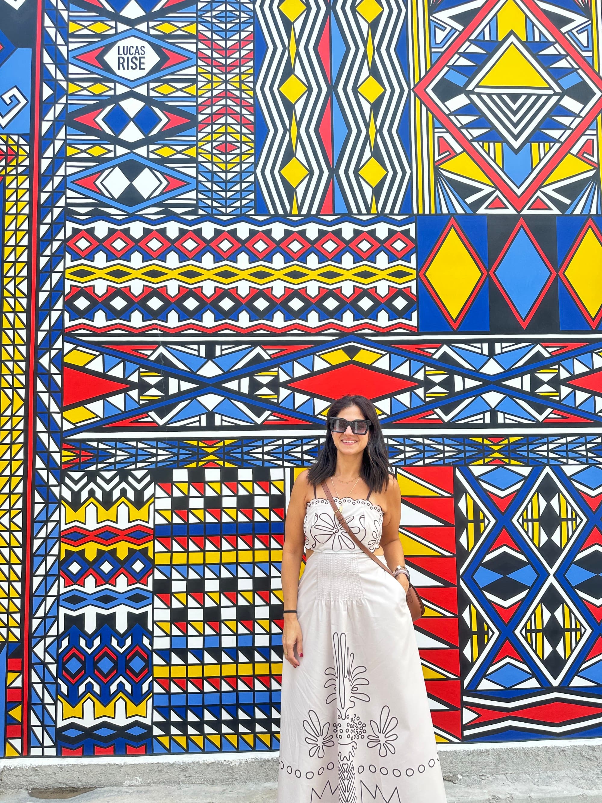 Woman standing in front of a colorful wall mural wearing a long white sundress and sunglasses.