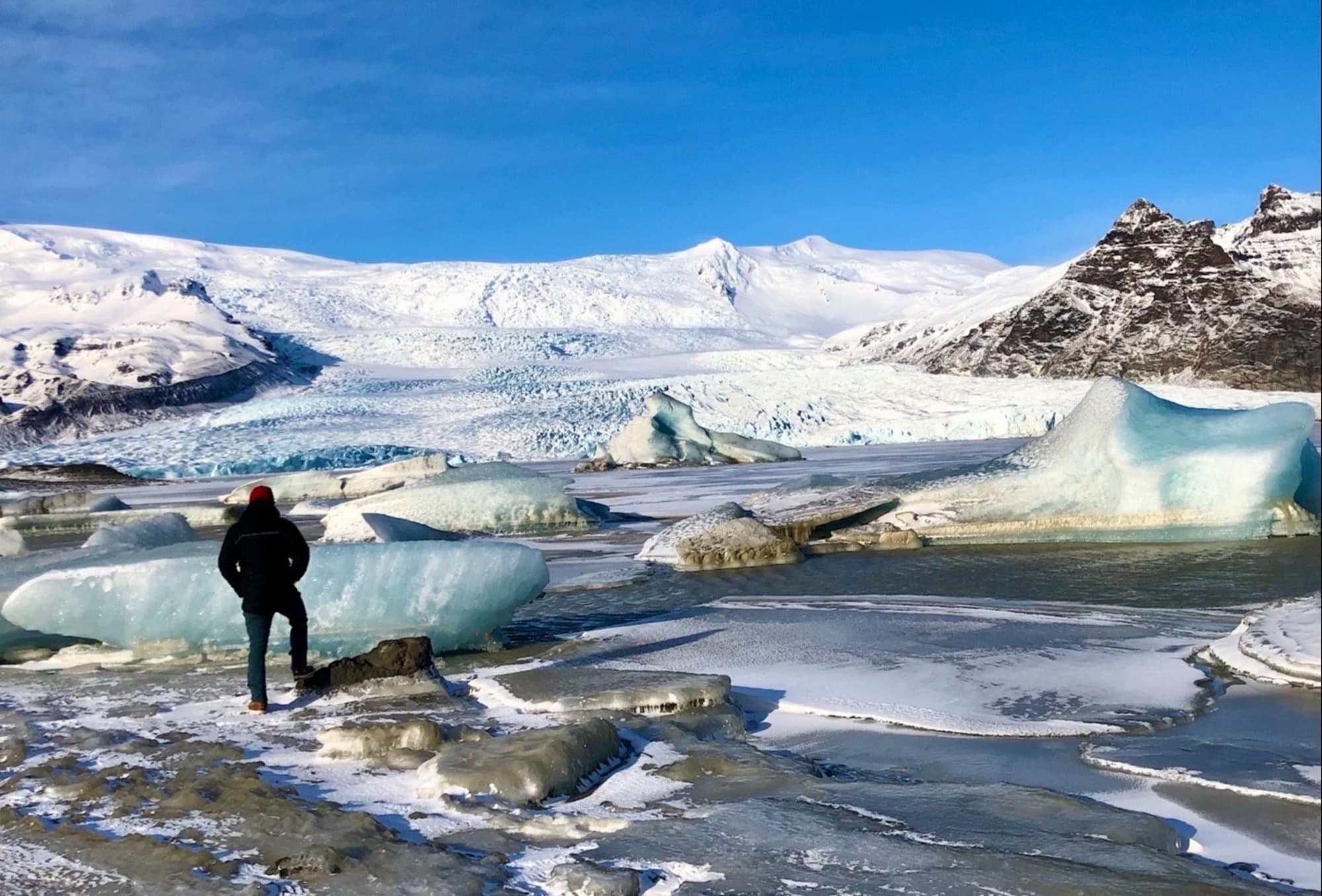 A lone explorer stands before a breathtaking glacial vista, dwarfed by the immense ice formations and glaciers under a pristine blue sky.