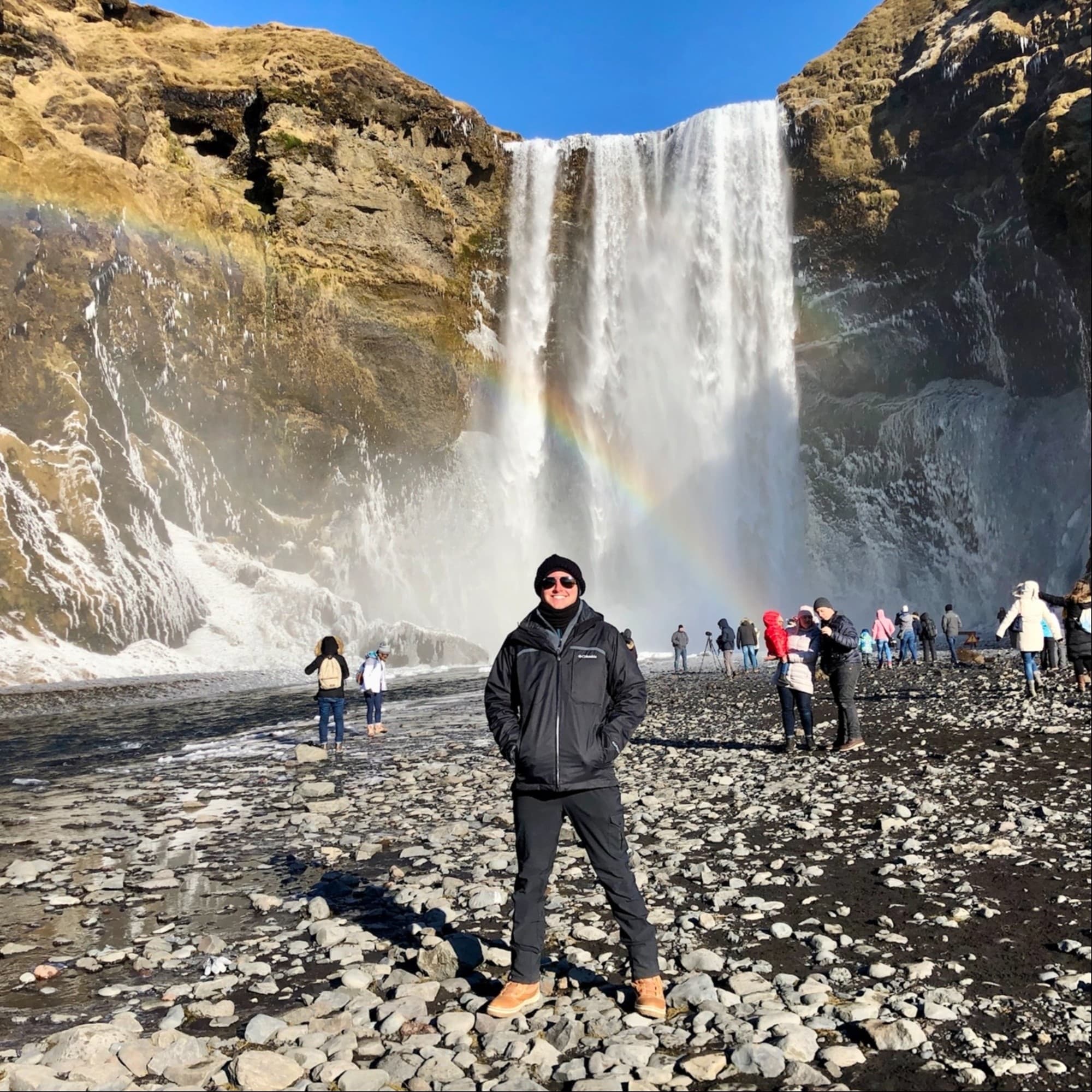 The image depicts a person standing in front of a waterfall with a rainbow during the daytime.