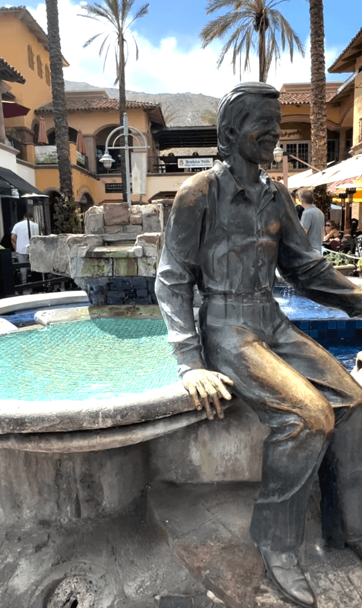 Grey sculpture of a man sitting on the side of a fountain.