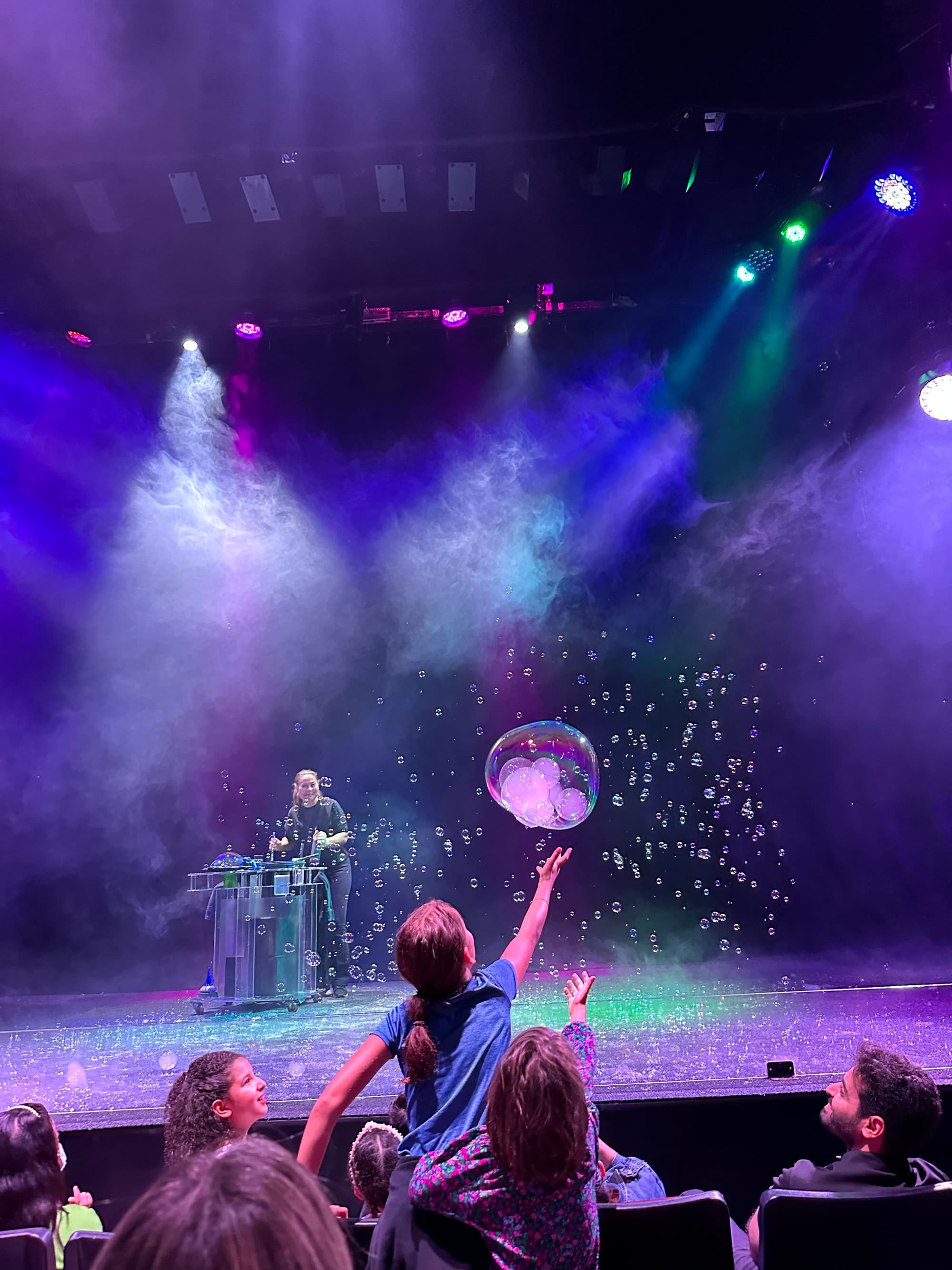 A child reaches out to a bubble during a vibrant bubble show, with an audience watching in the foreground.