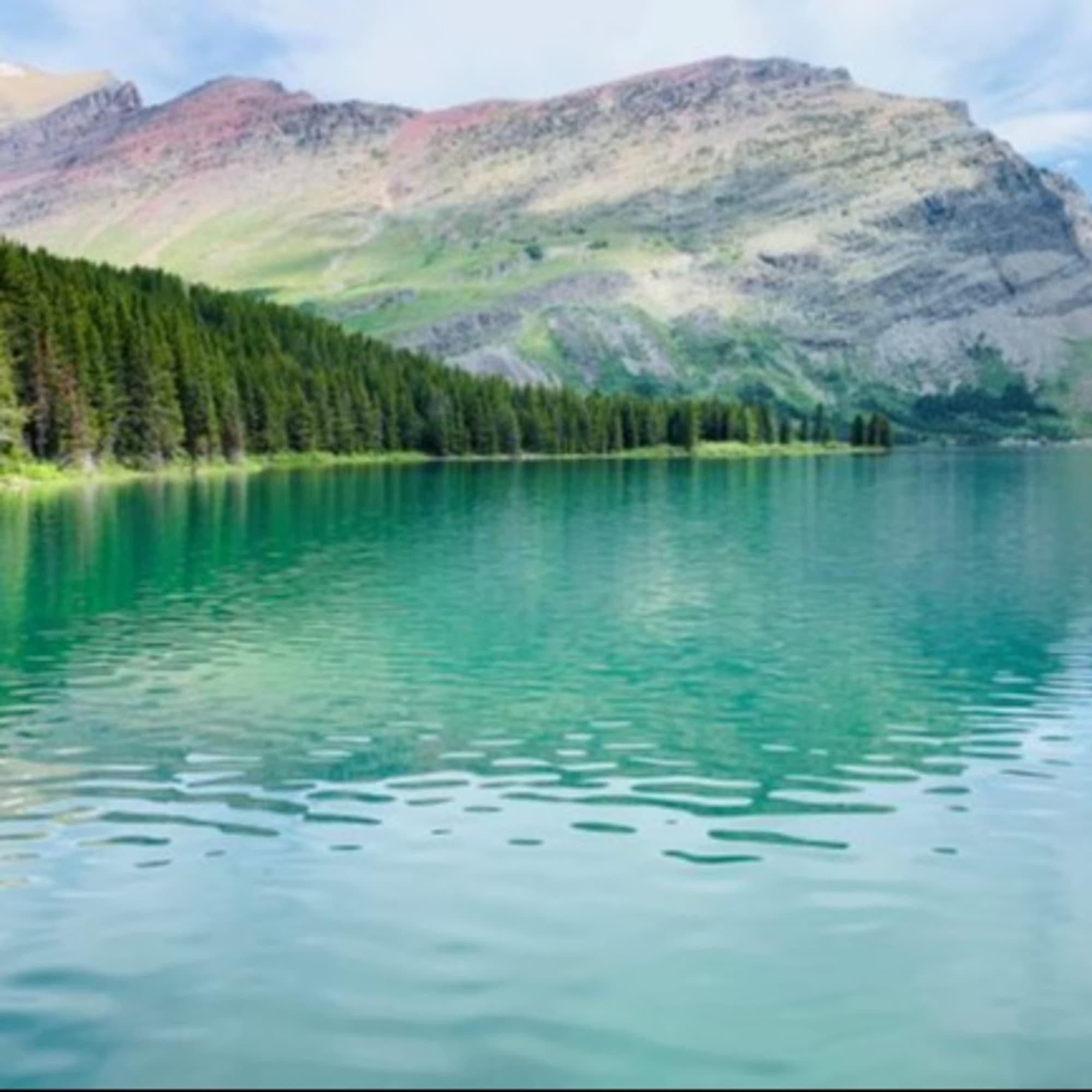 The image shows a serene lake with clear turquoise waters, surrounded by lush greenery and a rugged mountain backdrop under a blue sky.