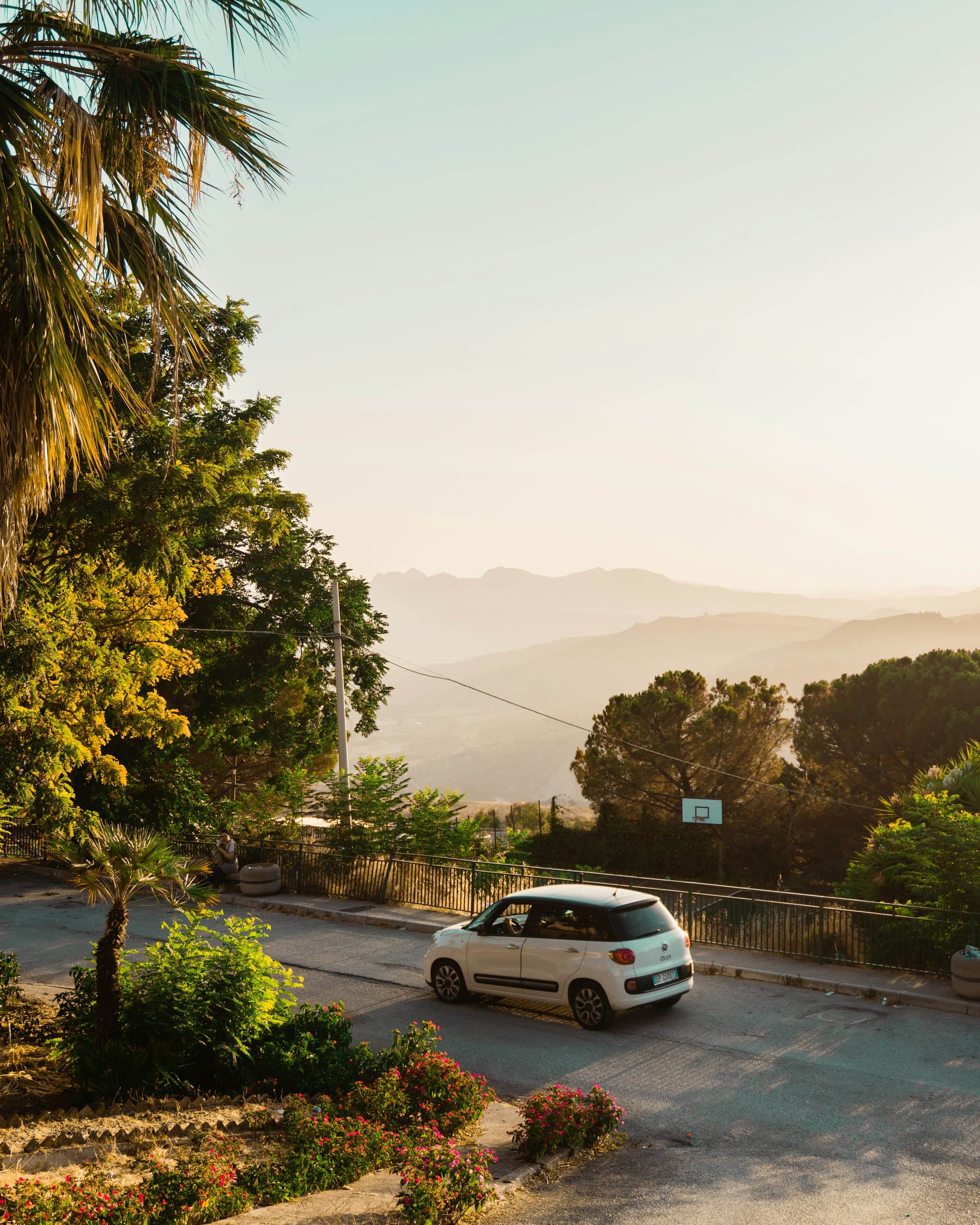A white car on the road surrounded by foliage with low mountains in the background.