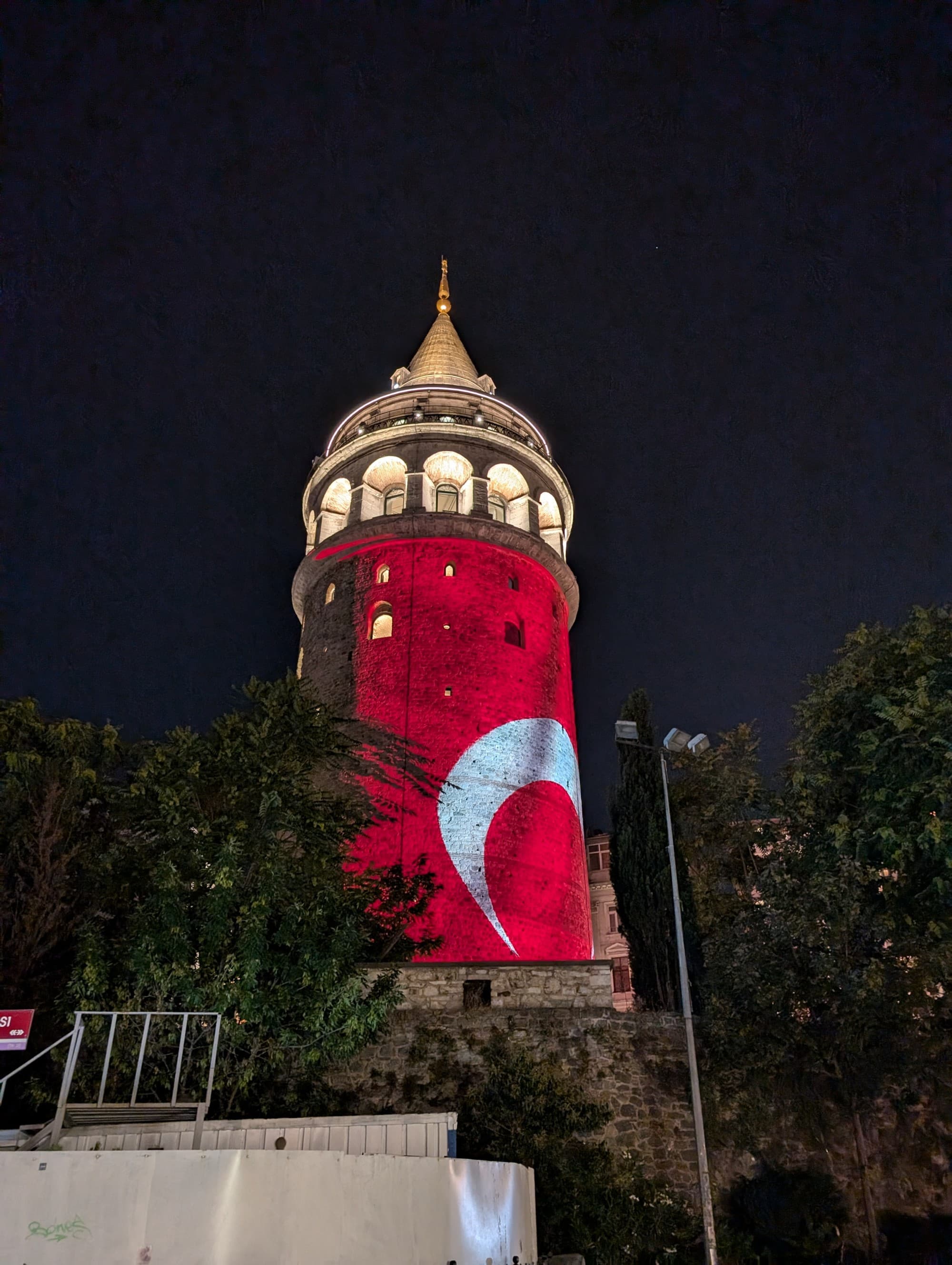 The image shows a tower illuminated at night with a projection of the Turkish flag, surrounded by foliage.