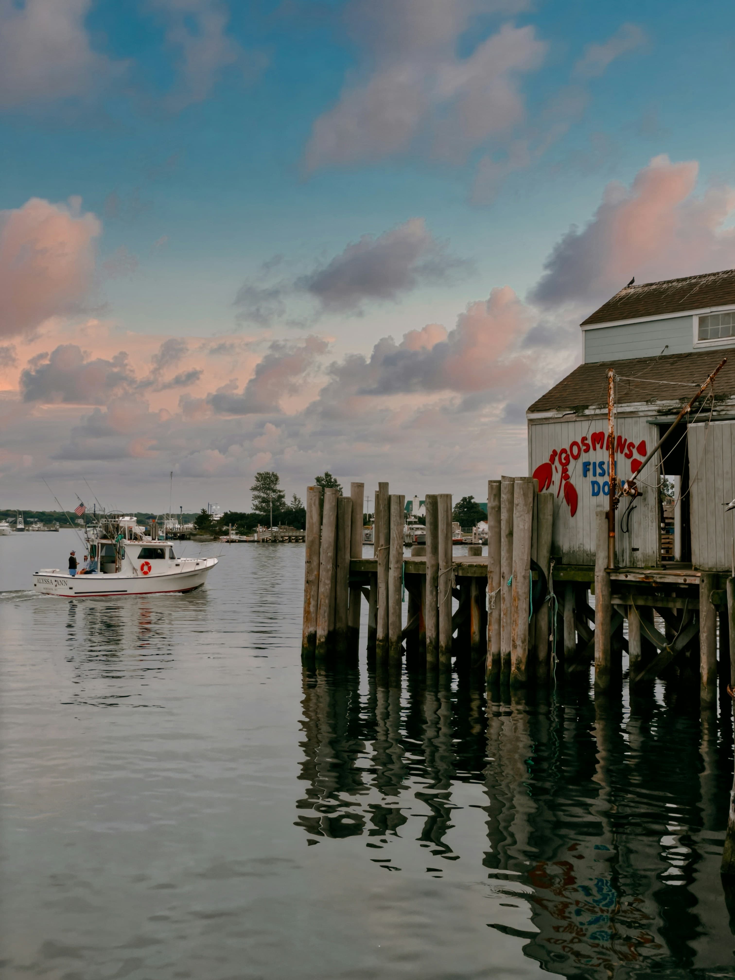 The image shows a pier with a white building with a red painted sign saying 'Gosmans', at sunset with pink fluffy clouds.