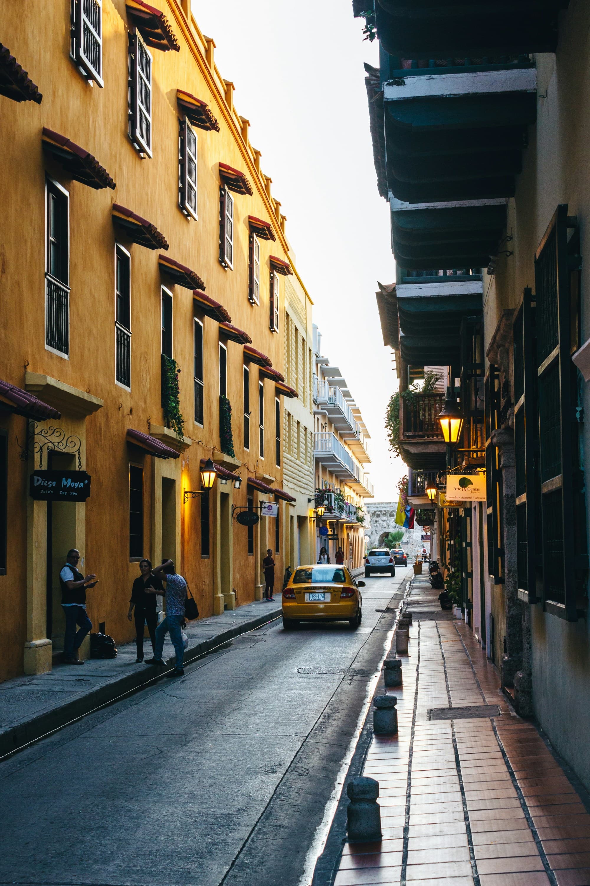 A view down a city street with people walking on the side walk and a car driving away, there is a yellow building on one side of the street.