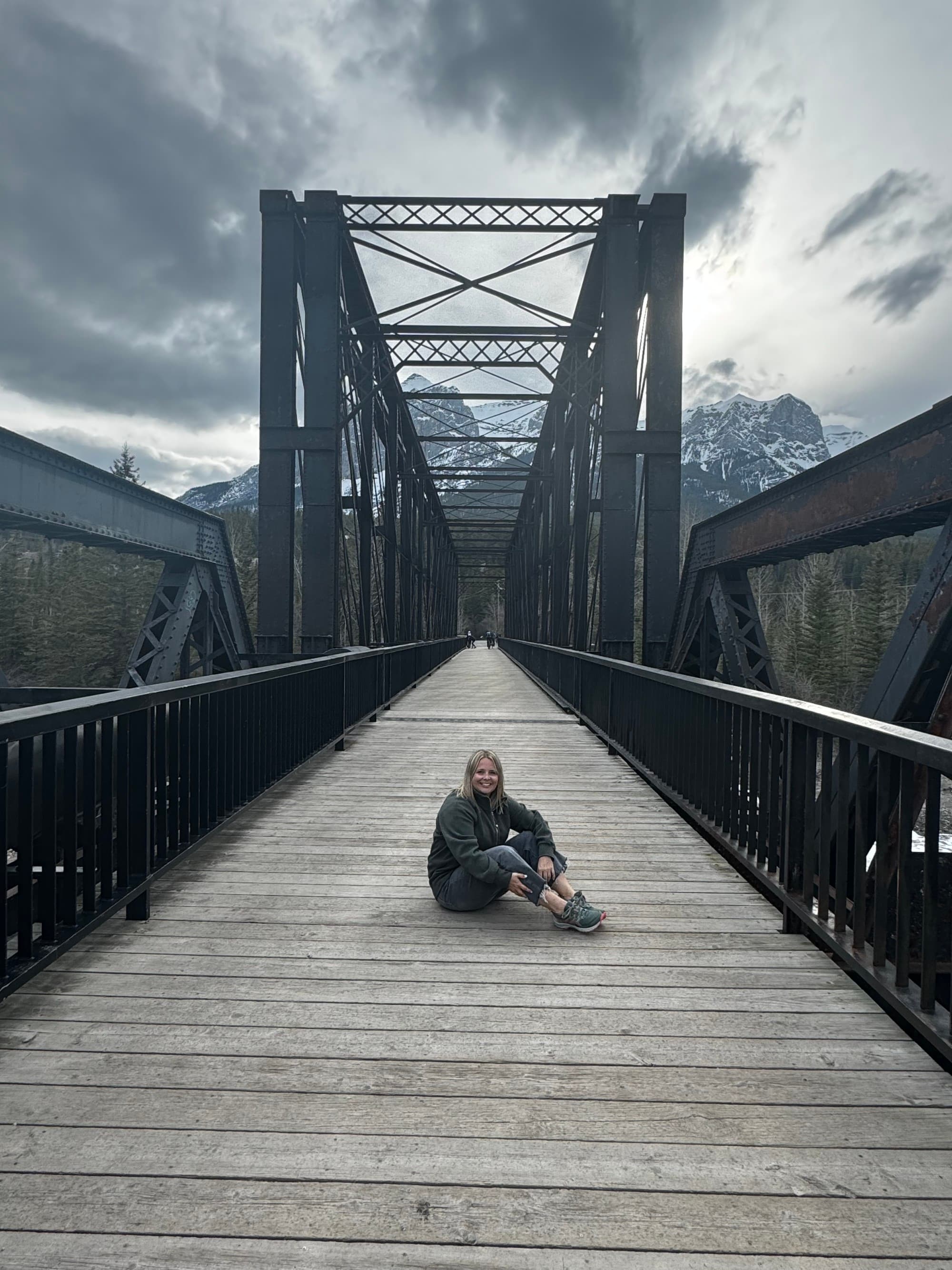 Travel advisor sitting on a wooden, pedestrian bridge with snow-covered mountains and gray skies in the background.