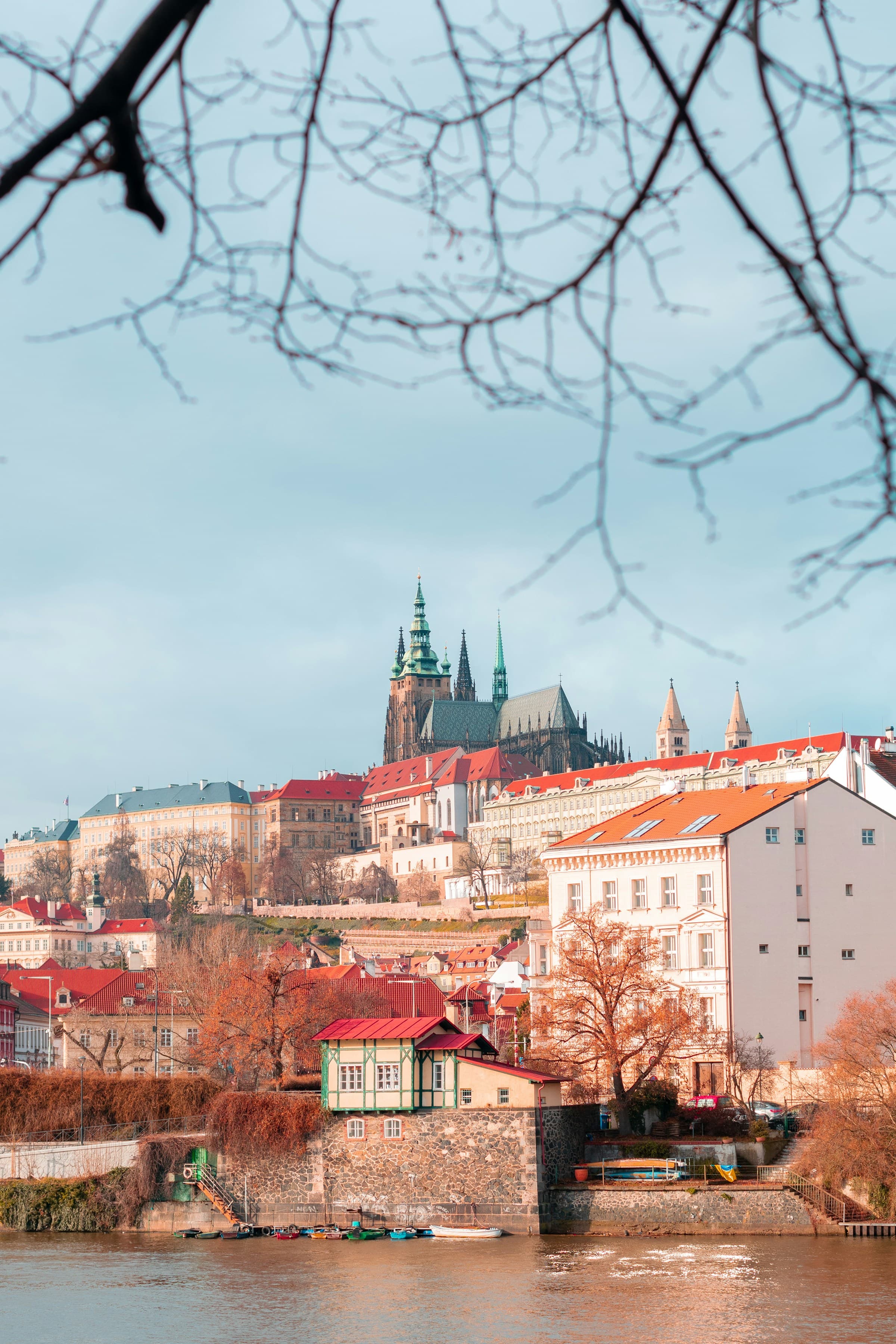 View of Prague Castle and white buildings with red roofs and autumn colored leaves from across a river.