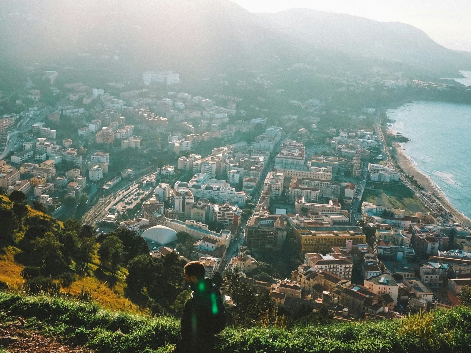A person hiking in Sicily on a grassy hill with the city, ocean and mountains in the background on a sunny day.