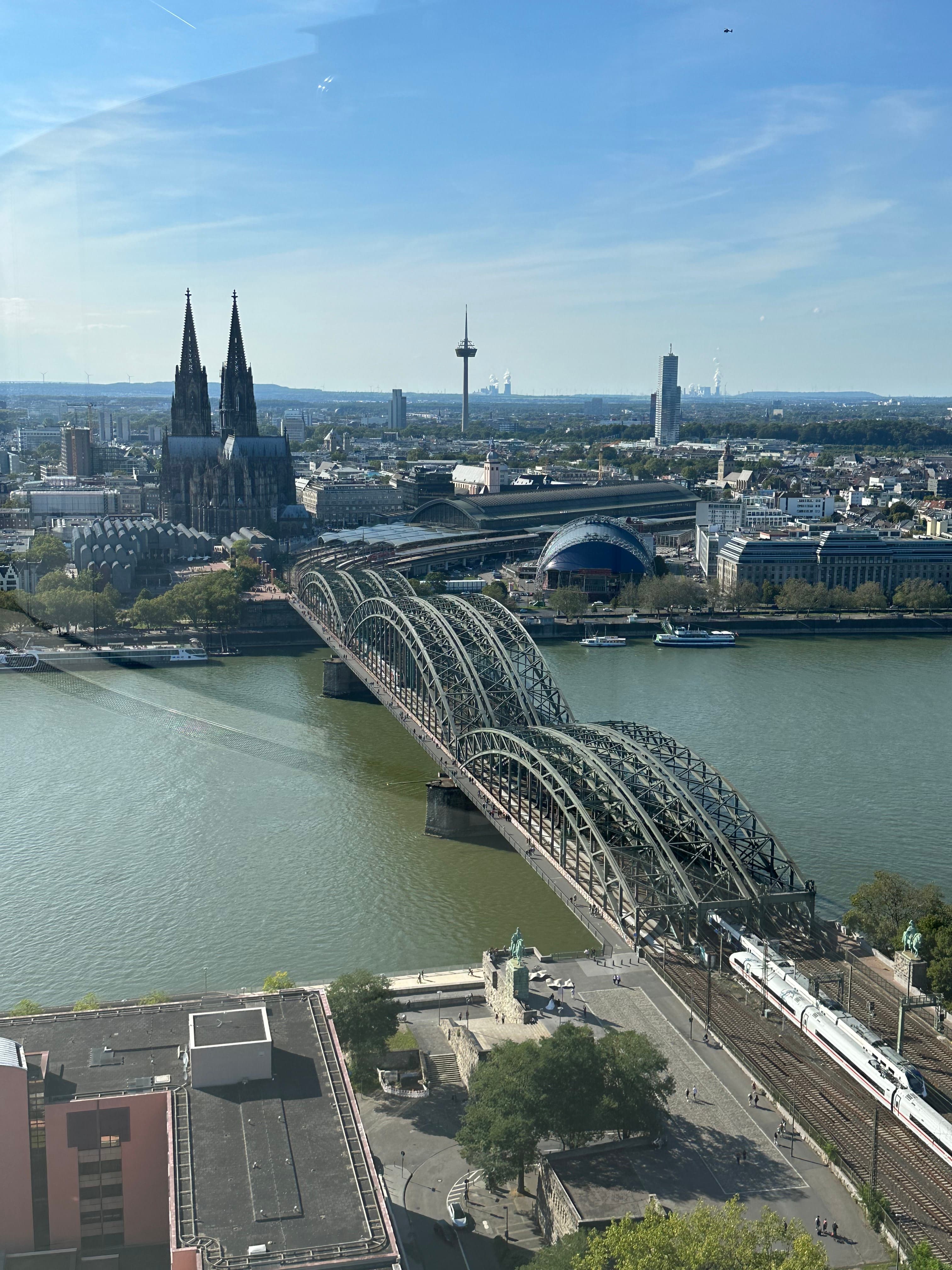 Aerial view of Cologne Triangle, a prominent landmark in Cologne, Germany. Picture features a bridge over a river with a cathedral and city skyline in the background on a clear day.