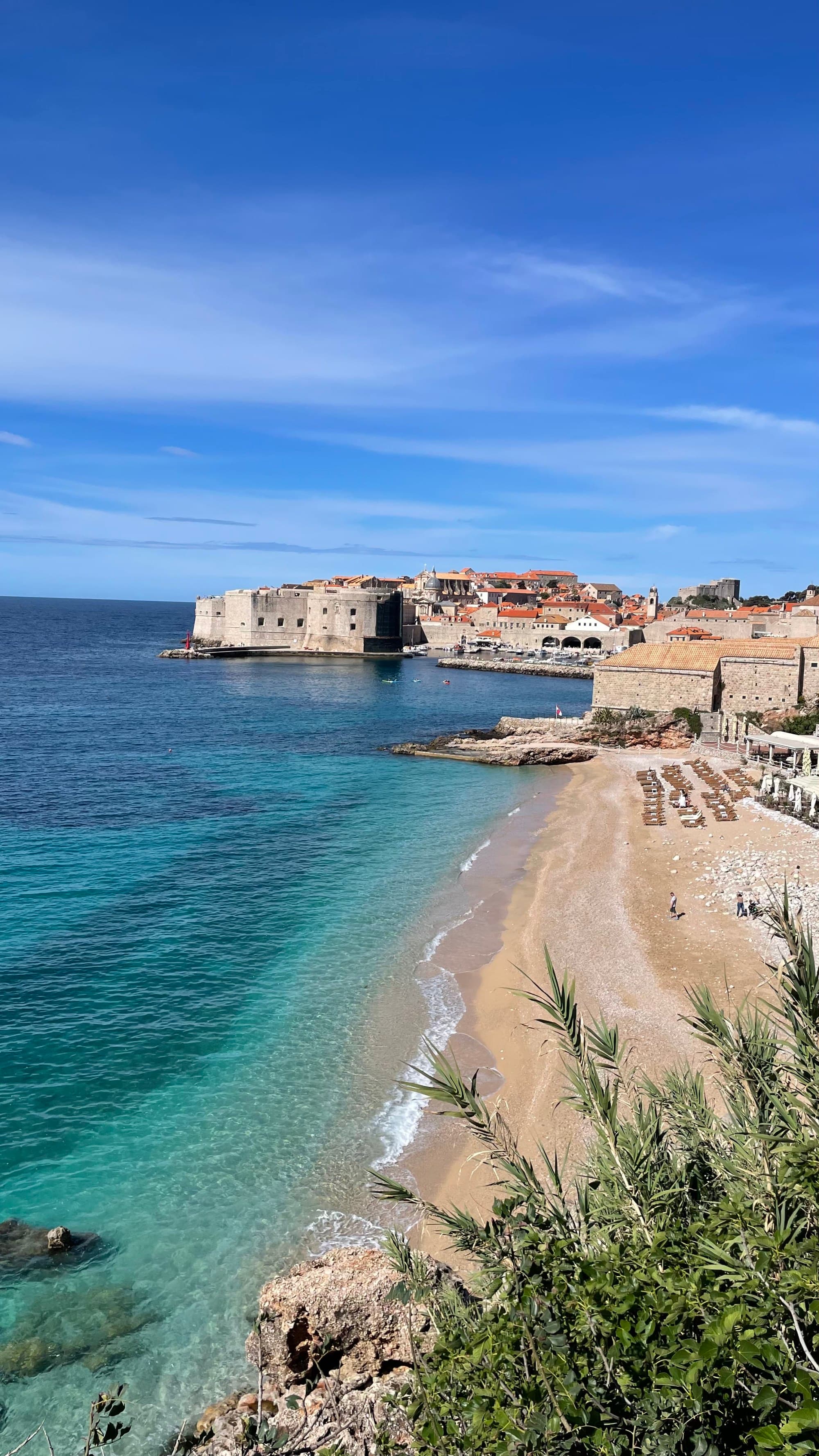 The image captures a picturesque coastal town in Croatia with historic architecture, clear blue waters and a sandy beach under a bright sky.