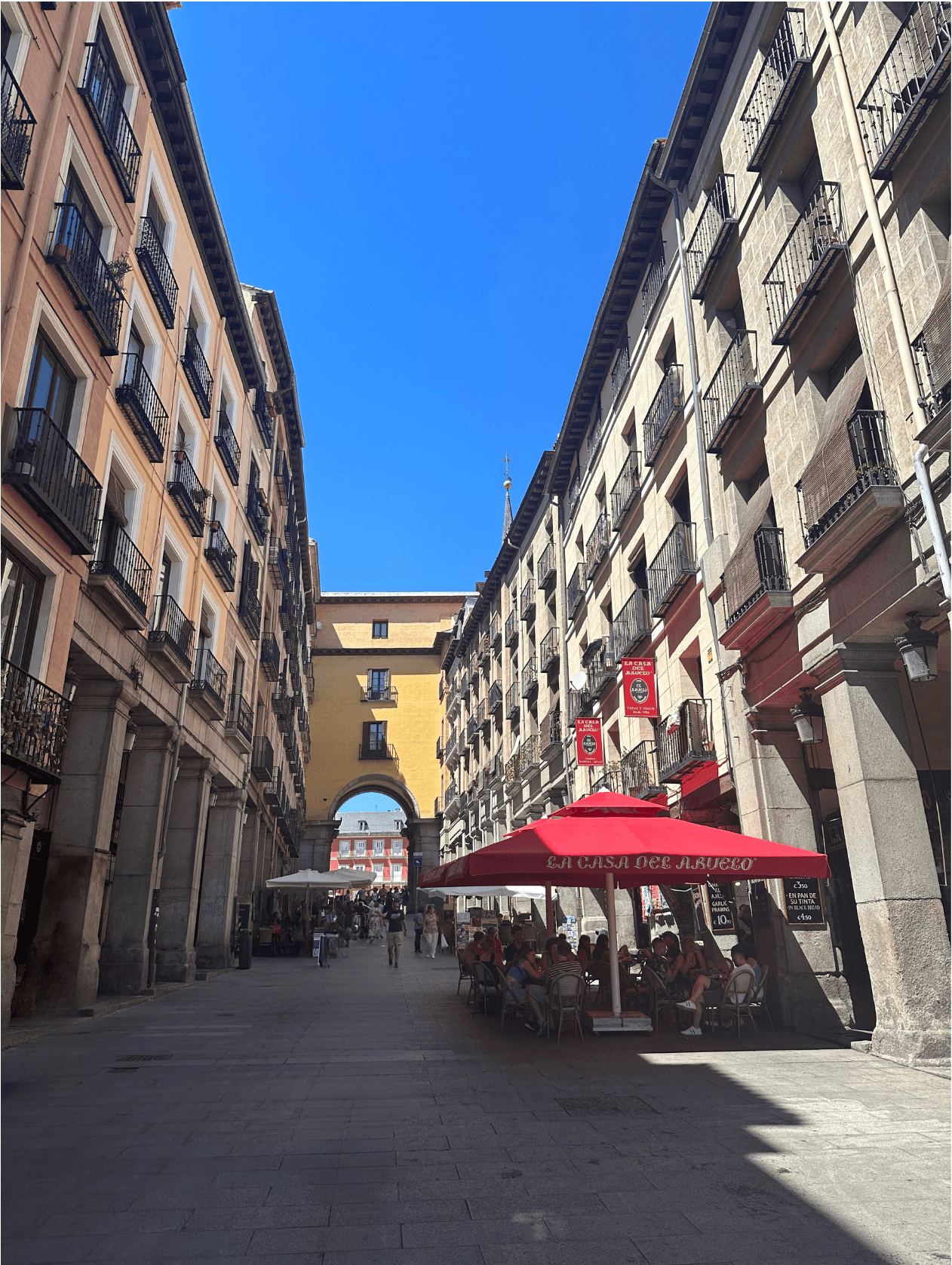 A European city street with a red umbrella, buildings with balconies on each side and a yellow archway in the distance.