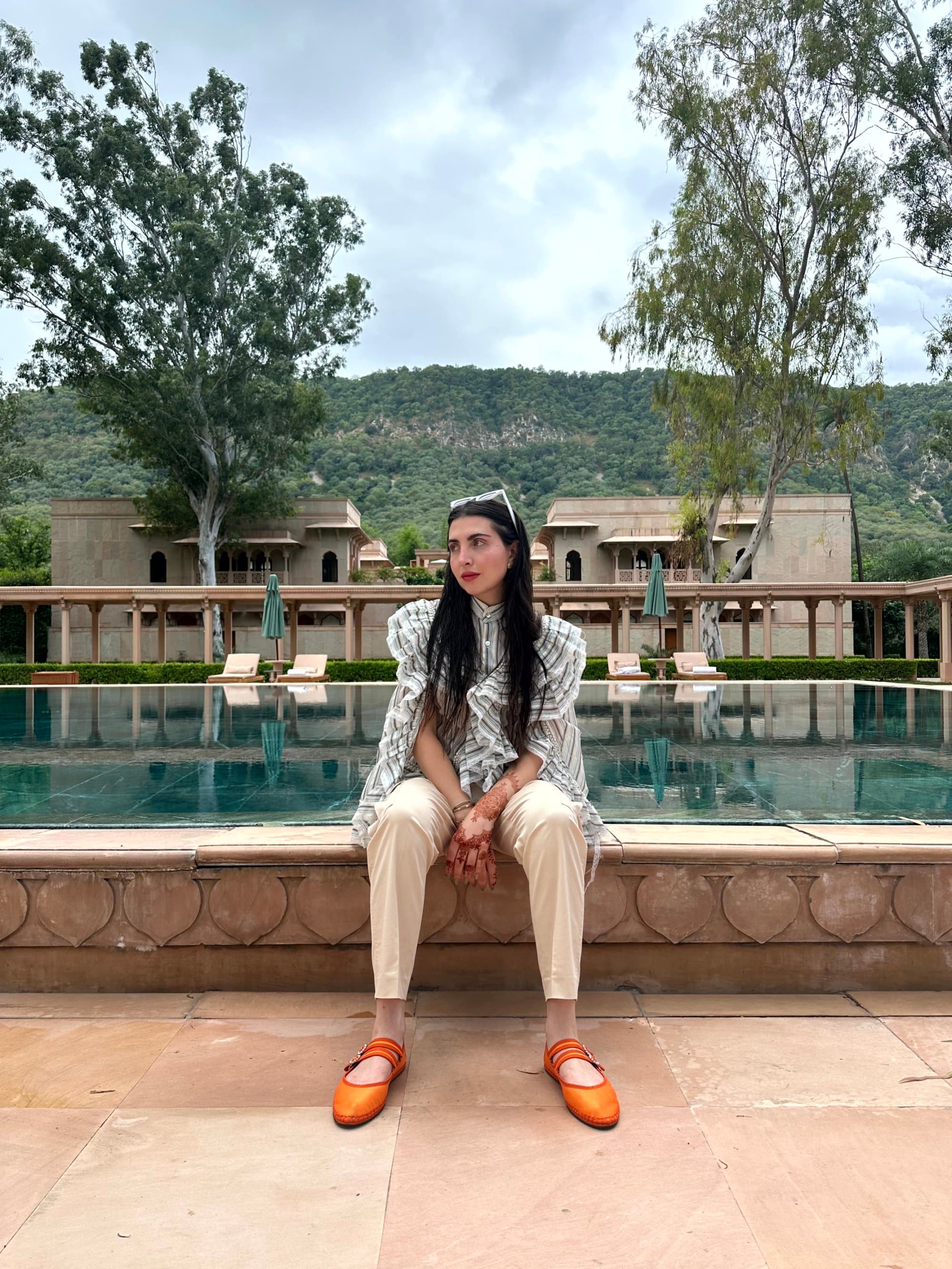 A woman wearing orange shoes, white shorts and a patterned blue top seated on a stone ledge in front of a swimming pool with trees and a mountain in the background.