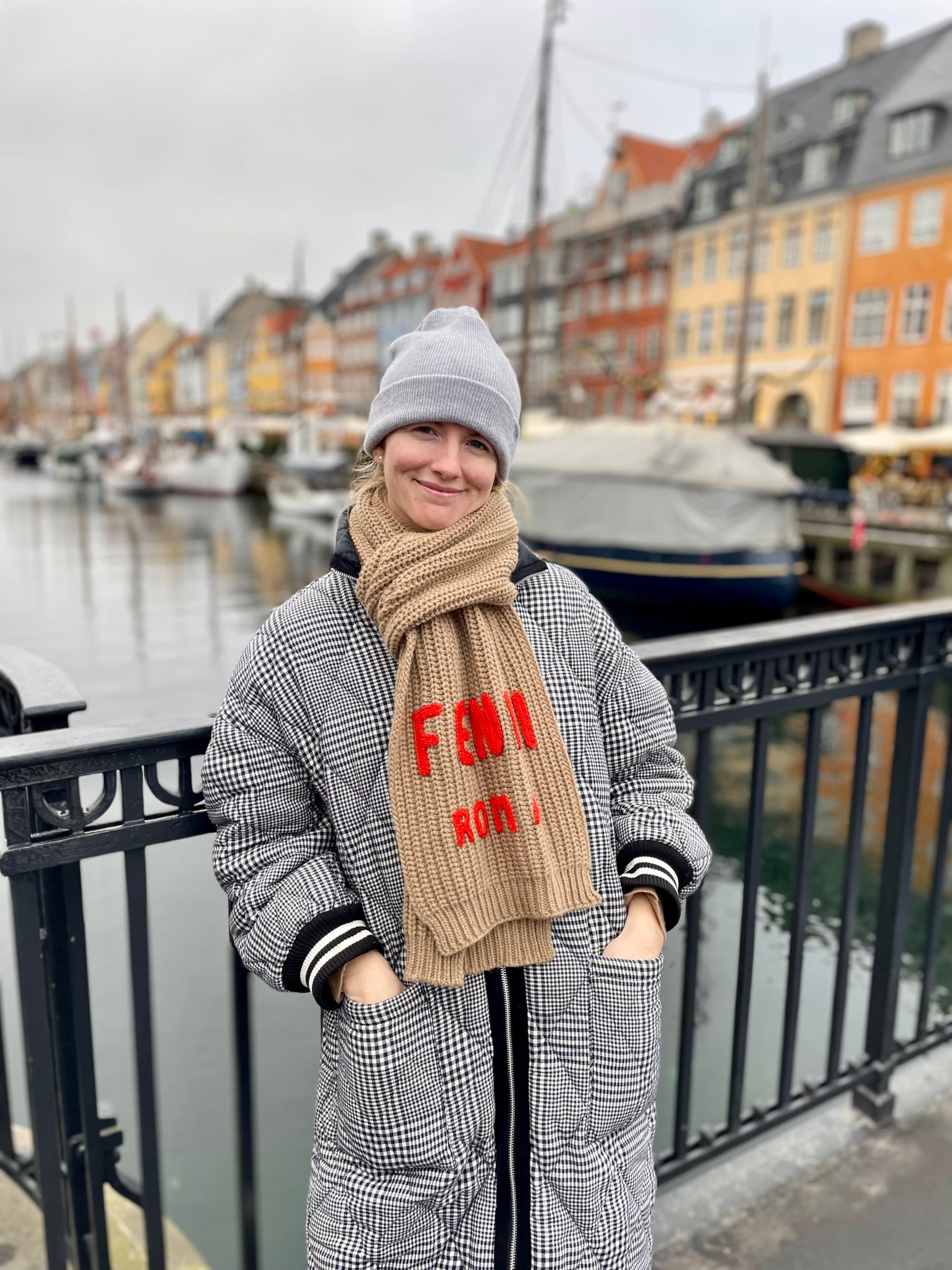 A picture of a girl in grey cap and coat posing on a bridge with colorful houses in the back during daytime.